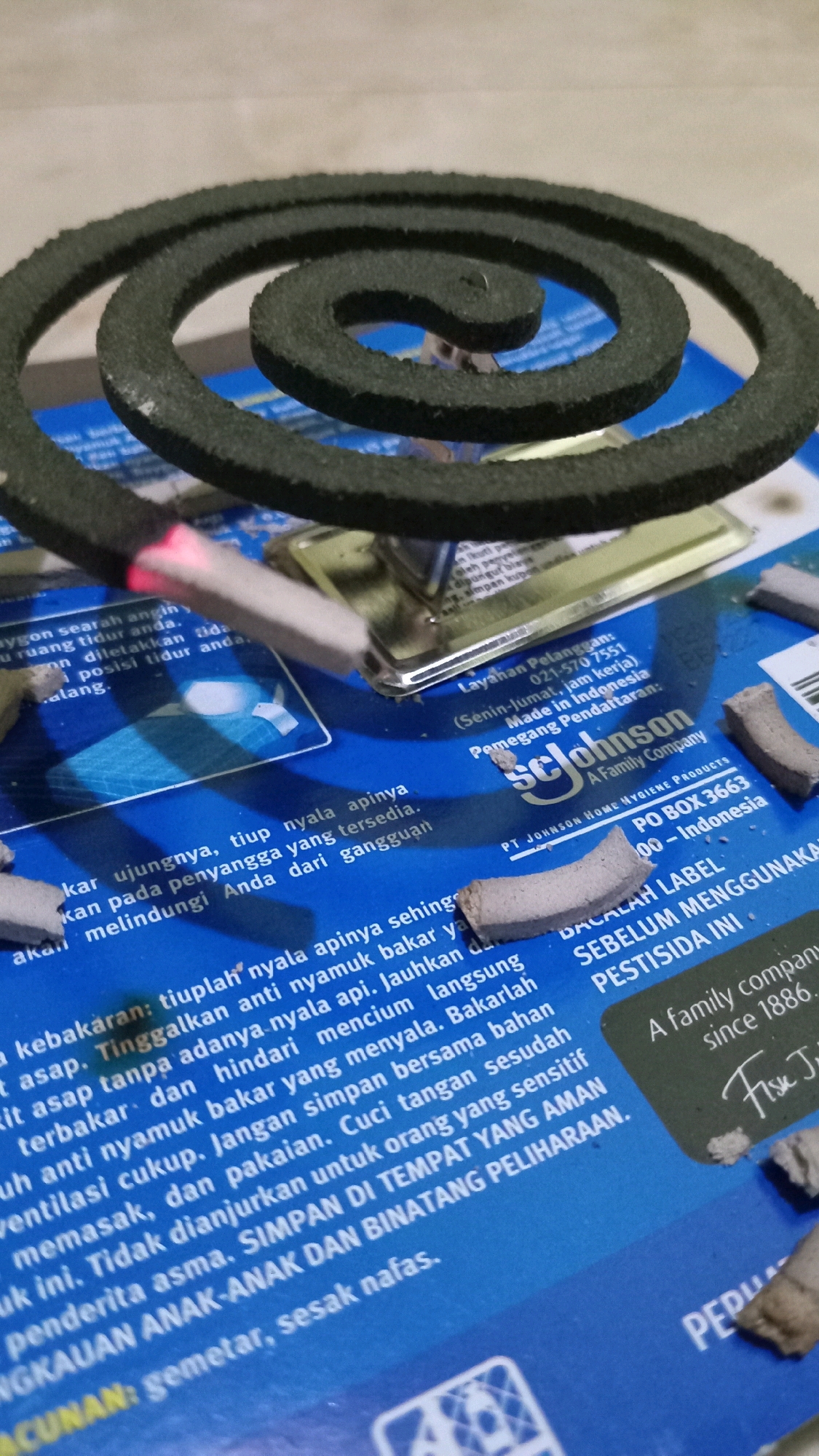 The image shows a mosquito coil, partially lit, with several broken pieces of mosquito coil scattered around it. The coil is dark green and is placed on a blue surface with text printed on it. The text appears to be in Indonesian and contains instructions and warnings related to the use of mosquito coils. Some of the text visible includes "Layahan Pelanggan:", "Made in Indonesia", "PT JOHNSON HOME HYGIENE PRODUCTS", "PO BOX 3663", "00 - Indonesia", "BACALAH LABEL SEBELUM MENGGUNAKAN PESTISIDA INI", "A family company since 1886.", "simpan di tempat yang aman untuk orang yang sensitif", "anak-anak dan binatang peliharaan.", and "gemetar, sesak nafas.". The image is taken from a slightly elevated angle, looking down at the mosquito coil and the surrounding debris. The overall environment appears to be an indoor setting, possibly a home. There are no people visible in the image. There is no information in the image to suggest the location in the city of Randuagung, Indonesia.