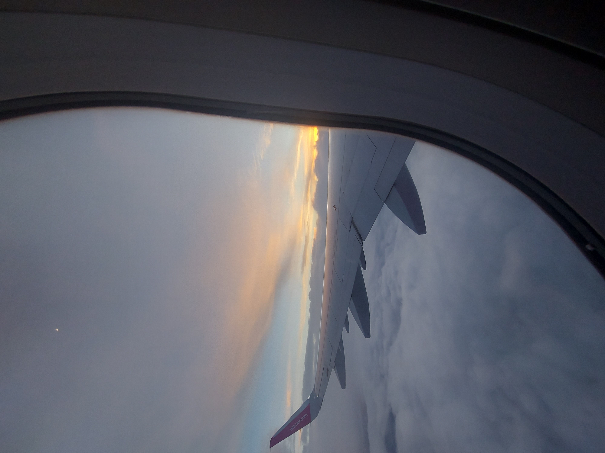 The image, taken from inside an airplane window, shows the wing of the plane and a view of the sky. The wing is white with a pink tip that reads "wizzair.com". The sky is a mix of light blue, orange, and gray, suggesting either sunrise or sunset. Below the wing, there are thick layers of clouds. The plane window frames the shot, adding a sense of depth. The image was not taken in Cherventsi, Bulgaria, and no additional details relating to people, objects, actions, interactions, or environment should be mentioned.