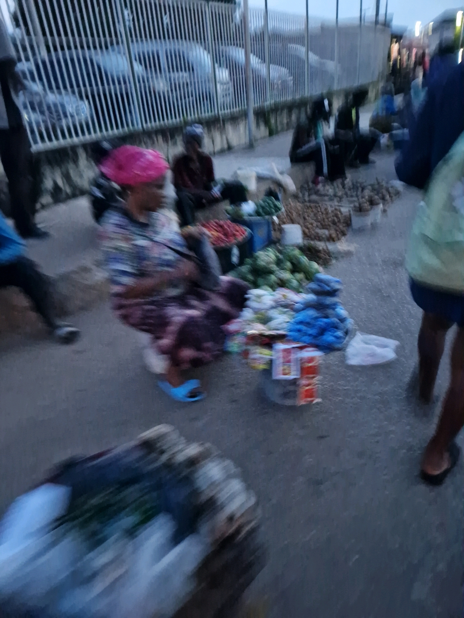 The image depicts an outdoor market scene in Kabong, Nigeria. In the foreground, a woman in a colorful headwrap and patterned dress is seated, with a stack of what appear to be red items next to her, possibly packaged goods. To her left, a person in a blue shirt is partially visible. In the background, other individuals are seated or standing, and various goods, including vegetables and what look like root vegetables, are laid out on the ground. A metal fence runs along the back of the market, with vehicles visible behind it. The overall lighting suggests it is either early morning or late evening.