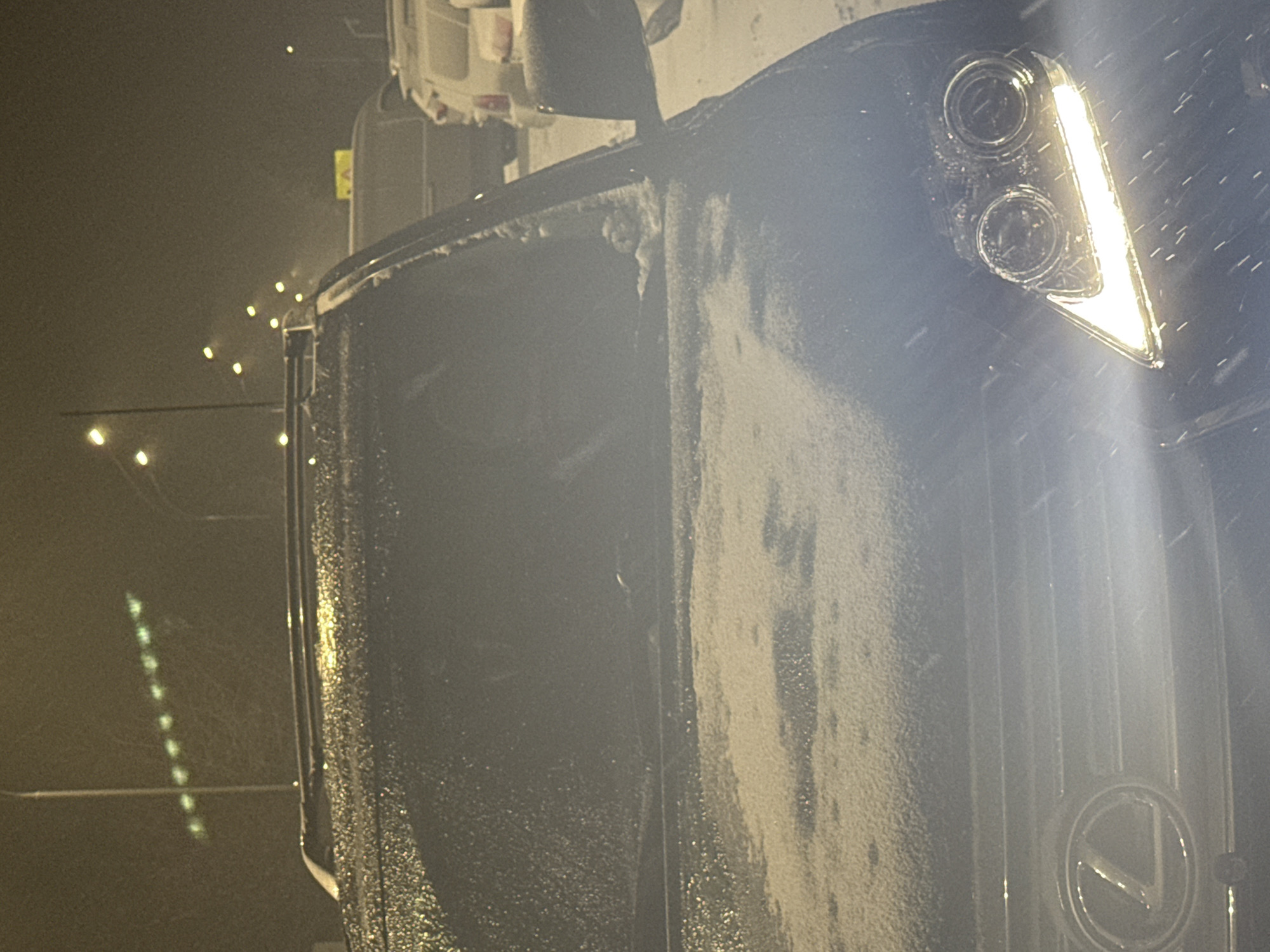 A close-up shot captures the front of a black Lexus vehicle in snowy conditions. The vehicle's headlight is illuminated, and the Lexus emblem is visible on the grill. The location is potentially Nizhnevartovsk, Russia, though no specific landmarks or details confirming this are directly visible in the image. The environment appears dark, with blurred lights suggesting a street scene at night. The vehicle is covered in snow or ice.