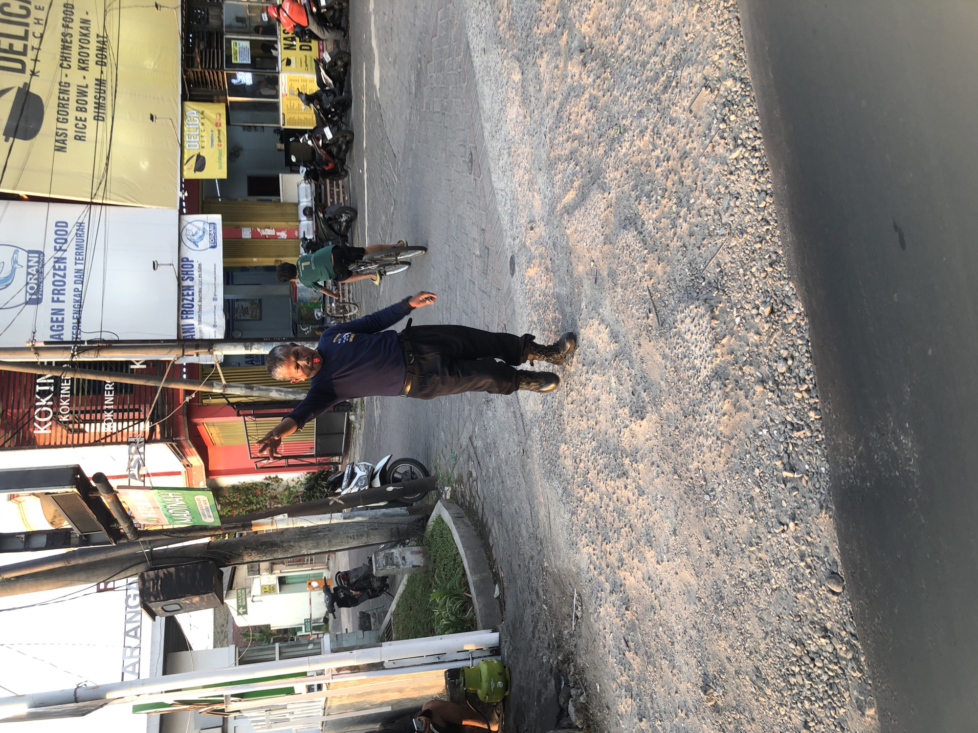 A man in a dark blue long-sleeved shirt and black pants, with a red object in his mouth, is standing in the middle of a street in Tenggulunan, Indonesia. He is gesturing with his left hand. The street is paved with cobblestones and gravel. Several motorcycles are parked on the right side of the street. Buildings with signs are visible in the background. One sign reads "TORANI FROZEN SHOP". Another sign reads "DELICA KITCHEN". The sun is setting, casting long shadows.