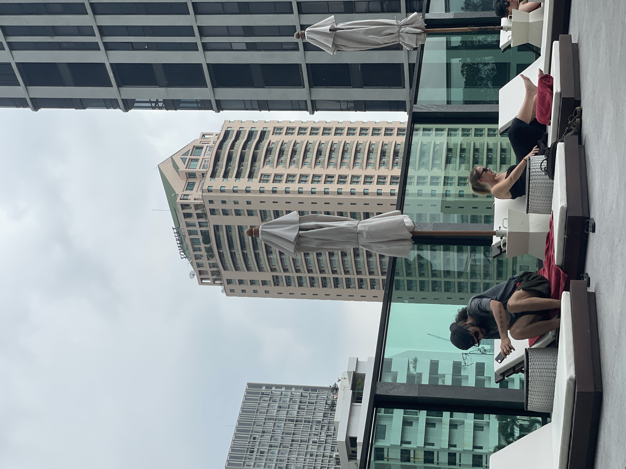 This image captures a contemporary urban scene, likely a rooftop or elevated terrace, featuring modern architecture and people relaxing. The primary subjects are two individuals lounging on outdoor furniture. One person, a woman, is seated with her legs up on a white lounge chair, wearing dark clothing and sunglasses, holding a bag or item next to her. A red towel is visible near her feet. Another person, a man with a beard and wearing a cap and sunglasses, is also seated, leaning forward and looking at a smartphone. He is wearing a dark t-shirt and shorts. Both individuals are partially obscured by their positions and the angle.
The setting appears to be a poolside or elevated seating area, indicated by white lounge chairs and folded white umbrellas. A glass railing separates the foreground from the background, reflecting parts of the city. Beyond the railing, a dense urban landscape unfolds under an overcast sky, characteristic of Bangkok's weather. Several tall buildings dominate the background. One prominent building is a light-colored, multi-story structure with numerous windows and balconies, featuring a distinctive stepped or curved facade. The name "SOMERSET" is partially visible on the side of this building, suggesting it might be a hotel or serviced apartment. Another large, grey building with a grid-like window pattern is also visible, and a glimpse of a third, dark-colored building with large rectangular windows forms the top of the frame. The overall impression is one of leisure amidst a bustling city backdrop.