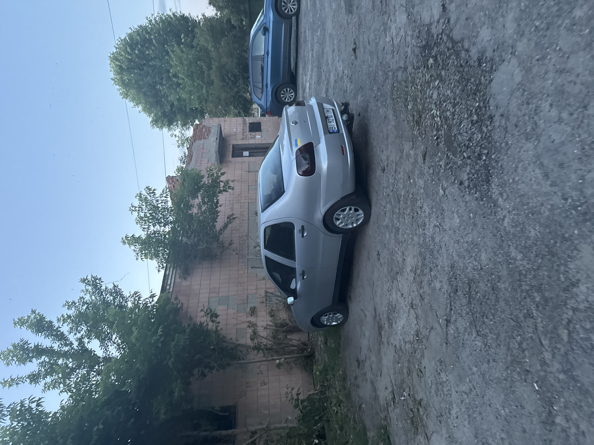 An outdoor, ground-level, eye-level medium shot captures a silver Fiat sedan parked on a gravel lot in front of a brick building in the city of Гожули, Ukraine. The building, partially obscured by trees, appears aged and has some graffiti on its walls. To the right of the Fiat, a blue SUV is also parked. The sky above is a clear blue, with a few dark objects that appear to be birds or insects. The scene is quiet and still, with no people present. The lighting suggests it is either early morning or late afternoon. The Fiat has a Ukrainian flag decal on its rear, and its license plate reads "BI 9210 IM".