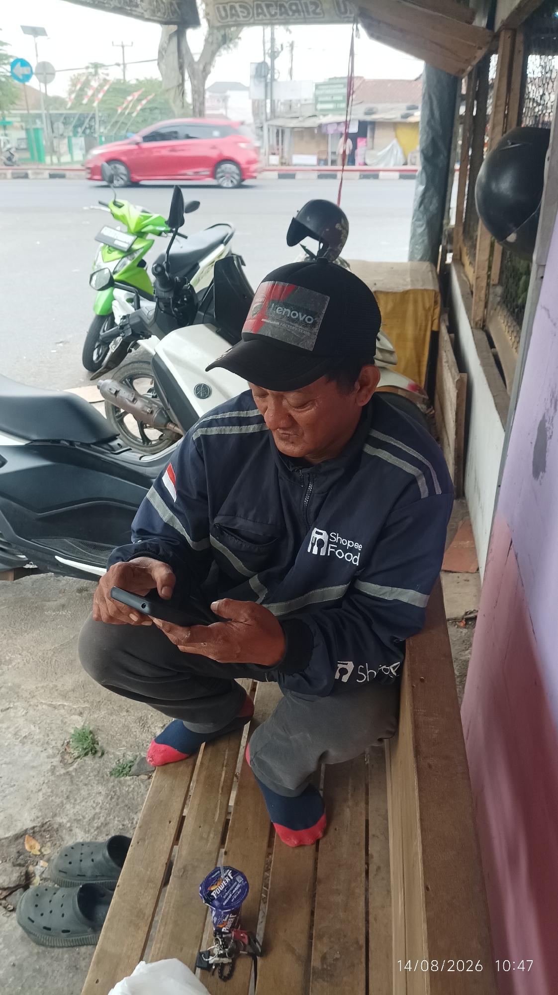 In Tasikmalaya, Indonesia, an Indonesian man wearing a dark blue jacket with "Shopee Food" branding and a black cap with a "Lenovo" logo sits on a wooden bench. He is holding and looking at a dark-colored smartphone in his hands. To his left, a green scooter is parked, and behind him, part of a white scooter is visible. The man is wearing grey pants and socks with red toes. A pair of grey clogs is on the ground to his left. On the bench next to him is a small, cylindrical object, possibly a can or container, with "POWER F" visible on its label, along with a keychain. The scene appears to be outdoors, with a road and buildings visible in the background. A red car is seen driving on the road. The date "14/08/2026" and time "10:47" are visible in the bottom right corner of the image.