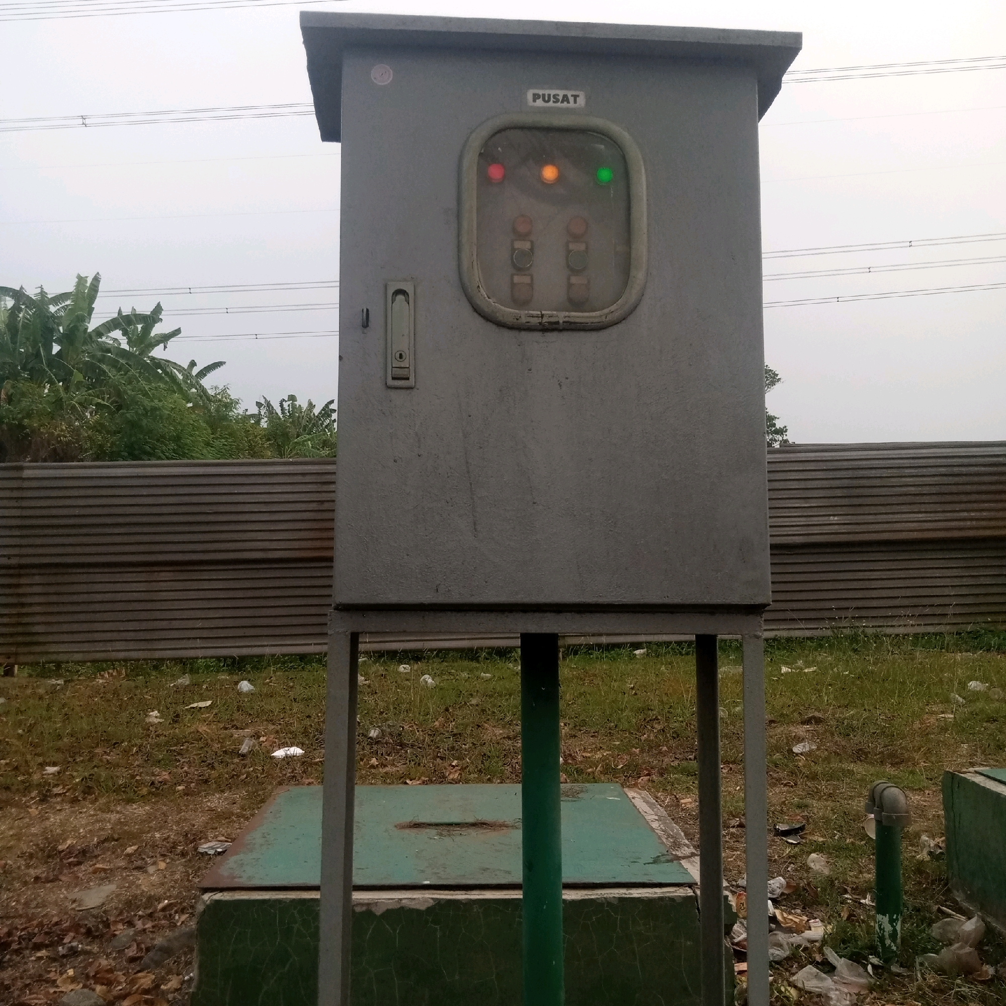 A grey electrical control box stands on four metal legs in an outdoor setting. The box has a single door with a silver latch on the left side. On the front of the box, a clear plastic-covered panel displays three illuminated indicator lights: red on the left, orange in the middle, and green on the right. Above this panel, the word "PUSAT" is printed. The box is situated in Cijeruk, Indonesia. Behind the box, a corrugated metal fence runs horizontally. In the background, lush green vegetation, including banana trees, is visible, and power lines are strung across the sky, which is overcast. The ground in front of the box is covered with dry grass, fallen leaves, and scattered litter. A green metal-covered box is partially visible on the ground directly in front of the electrical control box, supported by a dark green pole. To the right of the main subject, another green structure and a short, dark green pipe with a grey fitting are also present.