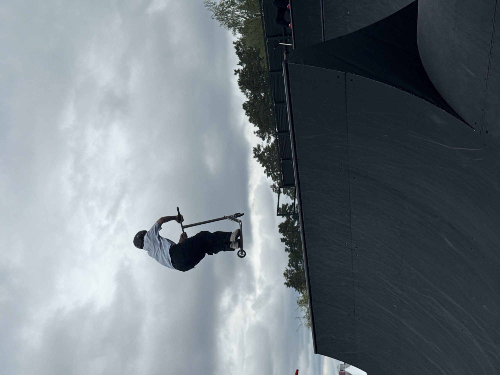 A person riding a scooter performs a trick against a backdrop of a cloudy sky and a dark, ramp-like structure. The person is mid-air, with the scooter positioned horizontally. They are wearing a helmet, a light-colored shirt, and dark pants. The ramp appears to be made of dark material. The environment suggests an outdoor skate park or similar recreational area. The location is in Yekaterinburg, Russia.