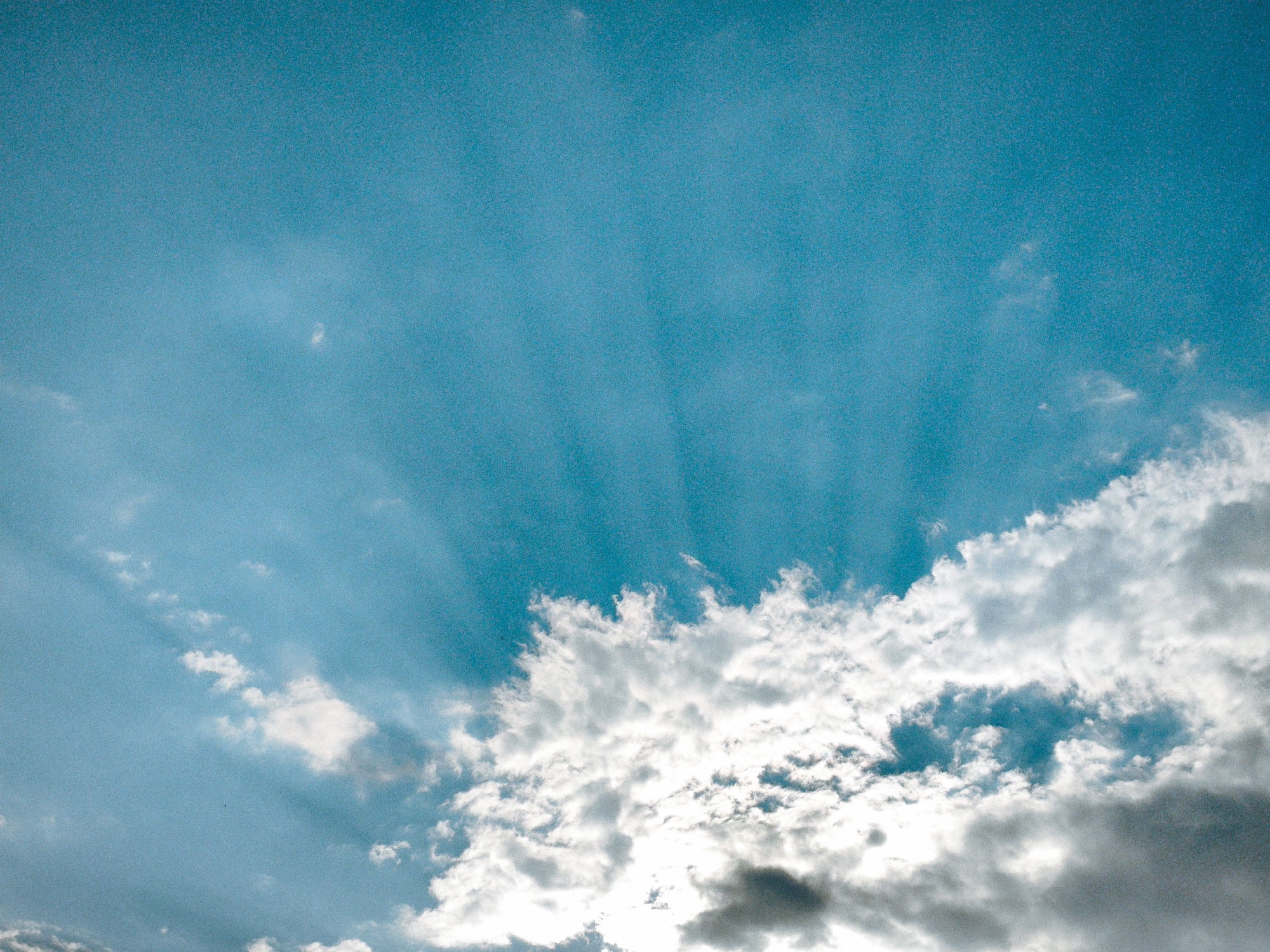The image is a low-angle shot of a bright, turquoise sky with scattered white and gray clouds. Sun rays are visible, piercing through the clouds in the upper left quadrant of the image. The clouds are dense and textured, with some areas appearing brightly illuminated by the sun and others casting shadows. The overall impression is a dramatic sky scene. The image was taken in an unknown city, Unknown.