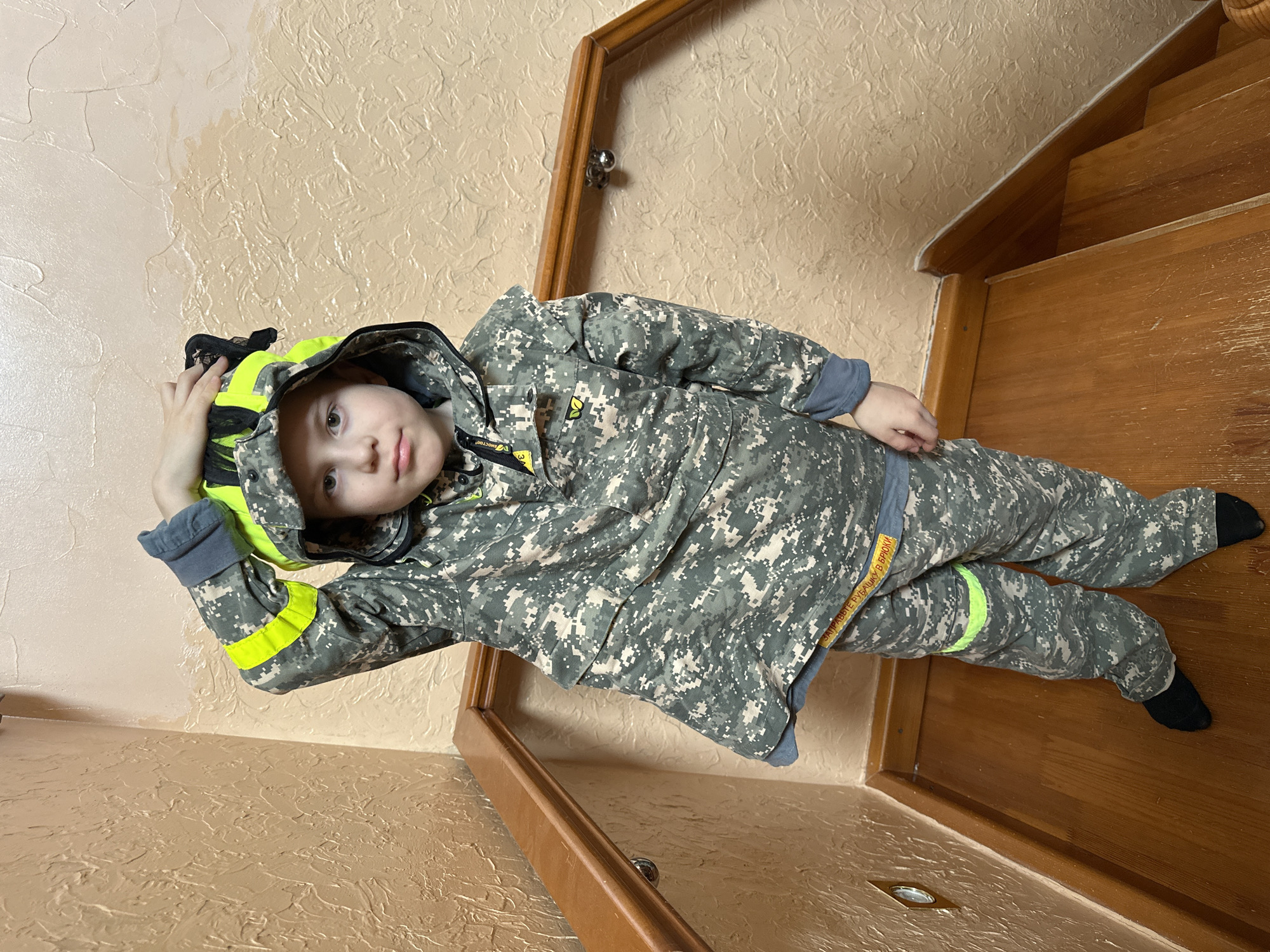 A young child dressed in camouflage patterned clothing with neon yellow accents is posing on a wooden staircase. The child is wearing a helmet with neon yellow trim. The background consists of textured beige walls and wooden handrails. The image appears to have been taken indoors. There is no explicit reference to Ilyinsky, Russia.