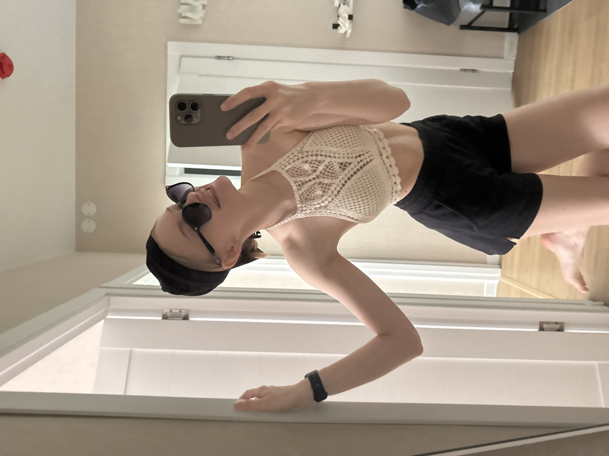 A young woman is taking a selfie in a mirror. She is wearing a white crochet halter top, black shorts, sunglasses, and a black cap. She is holding a phone with a triple-lens camera in her right hand. Her left arm is extended, with a black fitness tracker on her wrist. The background shows a white door and wall, and a glimpse of flooring. The photo appears to be taken in an indoor environment. There is no verifiable information indicating this photo was taken in Perm, Russia.