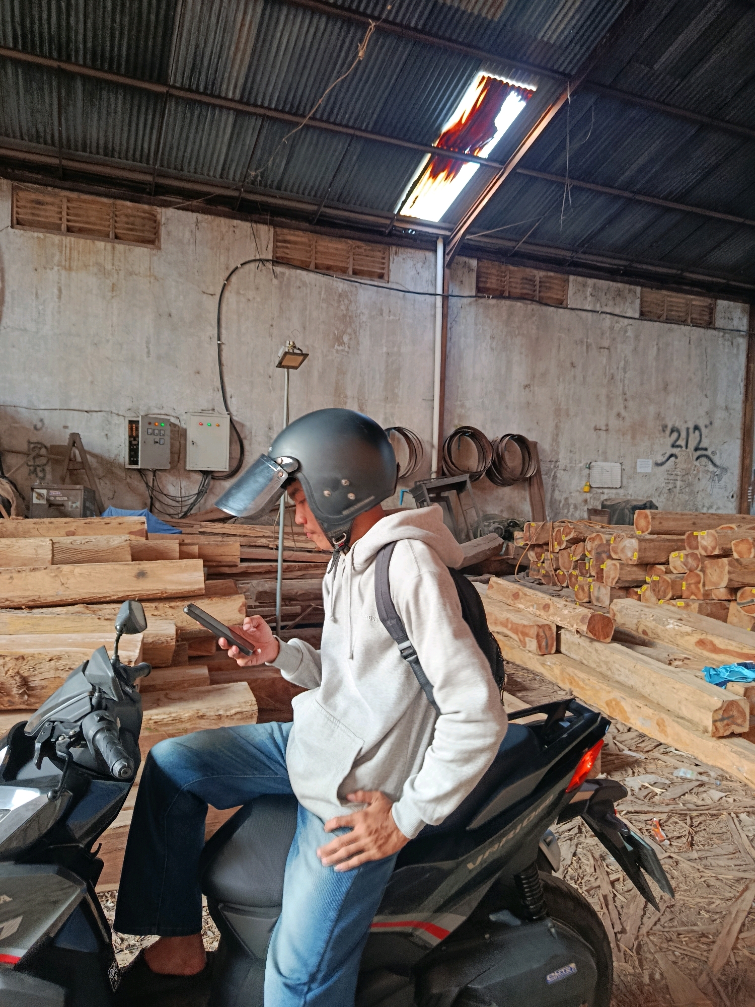 In a dimly lit warehouse in Sewon, Indonesia, a young person wearing a grey hoodie, blue jeans, and a dark helmet with a clear visor sits astride a black scooter. They are looking down at their phone, which they hold in their right hand. The scooter is parked amidst stacks of large, raw timber logs, some of which are bound with straps. The warehouse walls are rough concrete, and the corrugated metal roof has a large, broken section that allows sunlight to stream through, creating bright patches on the floor and illuminating dust motes in the air. Electrical conduits and junction boxes are visible on the walls, along with some graffiti. Several circular metal objects, possibly for binding logs, hang on the wall in the background. The overall impression is one of a working environment, perhaps a lumber yard or a woodworking shop.