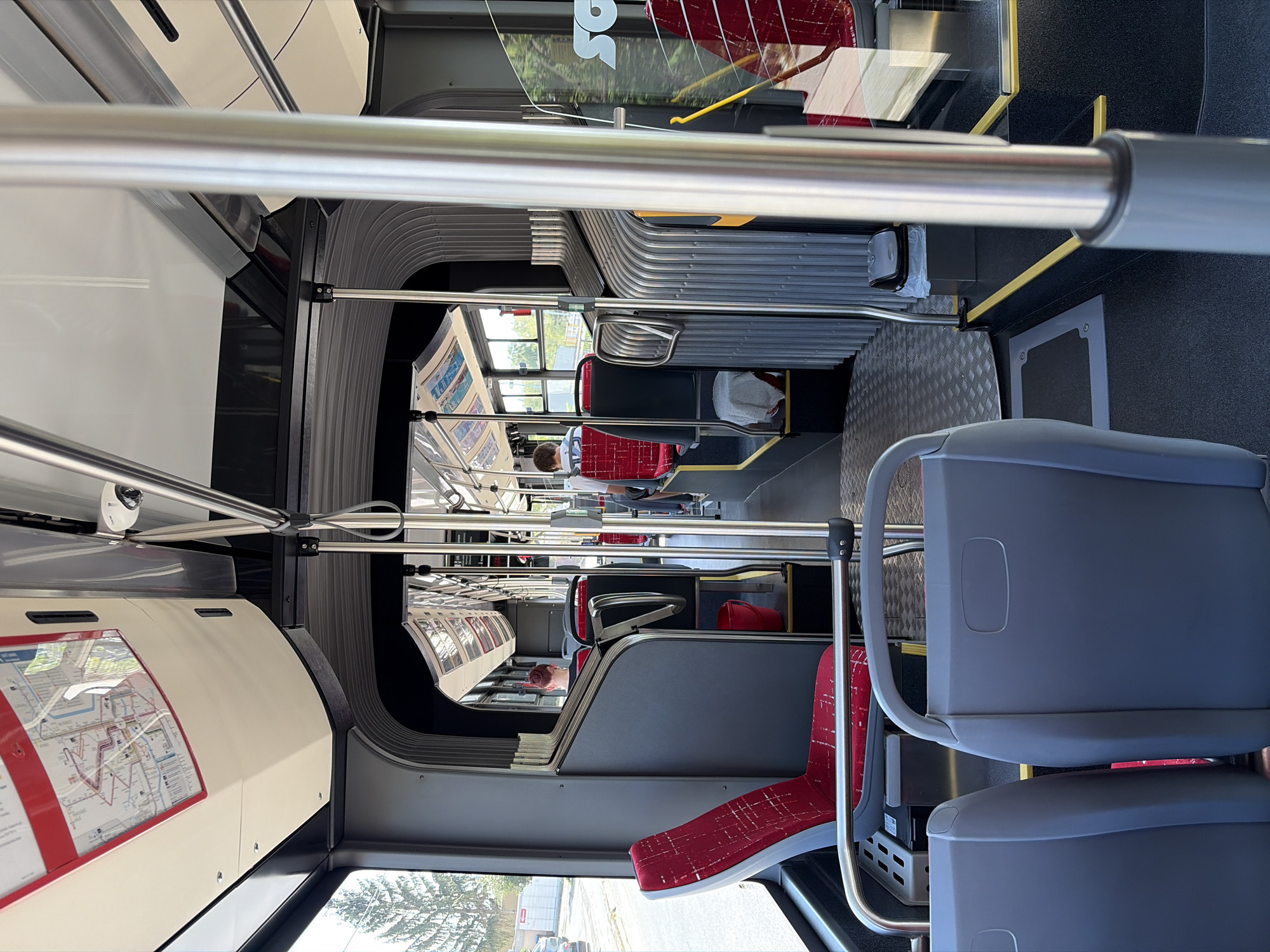 The image depicts the interior of a modern bus, likely a public transport vehicle, offering a perspective from one of the seats looking towards the rear. The bus features red and gray seating with visible safety poles and handrails. Overhead compartments and advertising or route maps are also present. The overall ambiance is one of a functional, well-maintained public transit environment. The presence of trees visible through the windows suggests an urban or suburban setting. Based on the provided context, this scene is located in the city of Nitra, Slovakia.