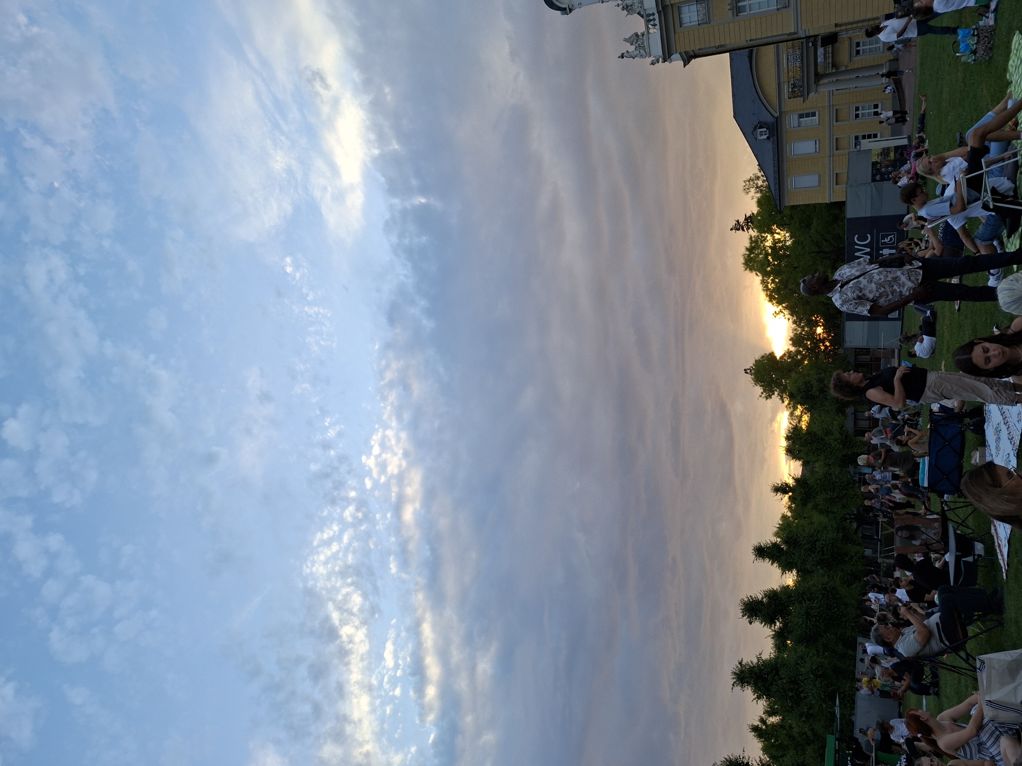 The image depicts an outdoor event at the Schlossgarten in Karlsruhe, Germany, during a sunset. A large crowd of people are gathered on a grassy lawn, seated on blankets and chairs, facing towards the right side of the frame. The sky is filled with clouds, with the setting sun casting a warm glow towards the horizon. In the background, a yellow building with classical architectural elements, likely part of the Karlsruhe Palace complex, is visible. Trees form a dense line behind the crowd. Some individuals are standing and interacting, while others are relaxed and seated, enjoying the event. The atmosphere appears casual and communal.