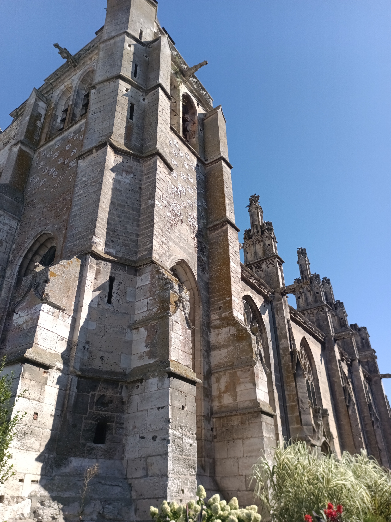 The image shows the exterior of a large, old stone church with a clear blue sky as the background. The church is constructed of large, weathered stone blocks, with visible signs of age and some crumbling. The architecture features Gothic elements, including pointed arches, intricate stone carvings, and gargoyles perched on the upper levels of the towers. Sunlight casts distinct shadows on the facade, highlighting the textures and architectural details. In the foreground, lush green foliage and some red flowers are visible, suggesting the church is surrounded by vegetation. The perspective is from a low angle, looking upwards at the imposing structure. This is the Church of Saint-Thomas-Becket in Le Neubourg, France.
