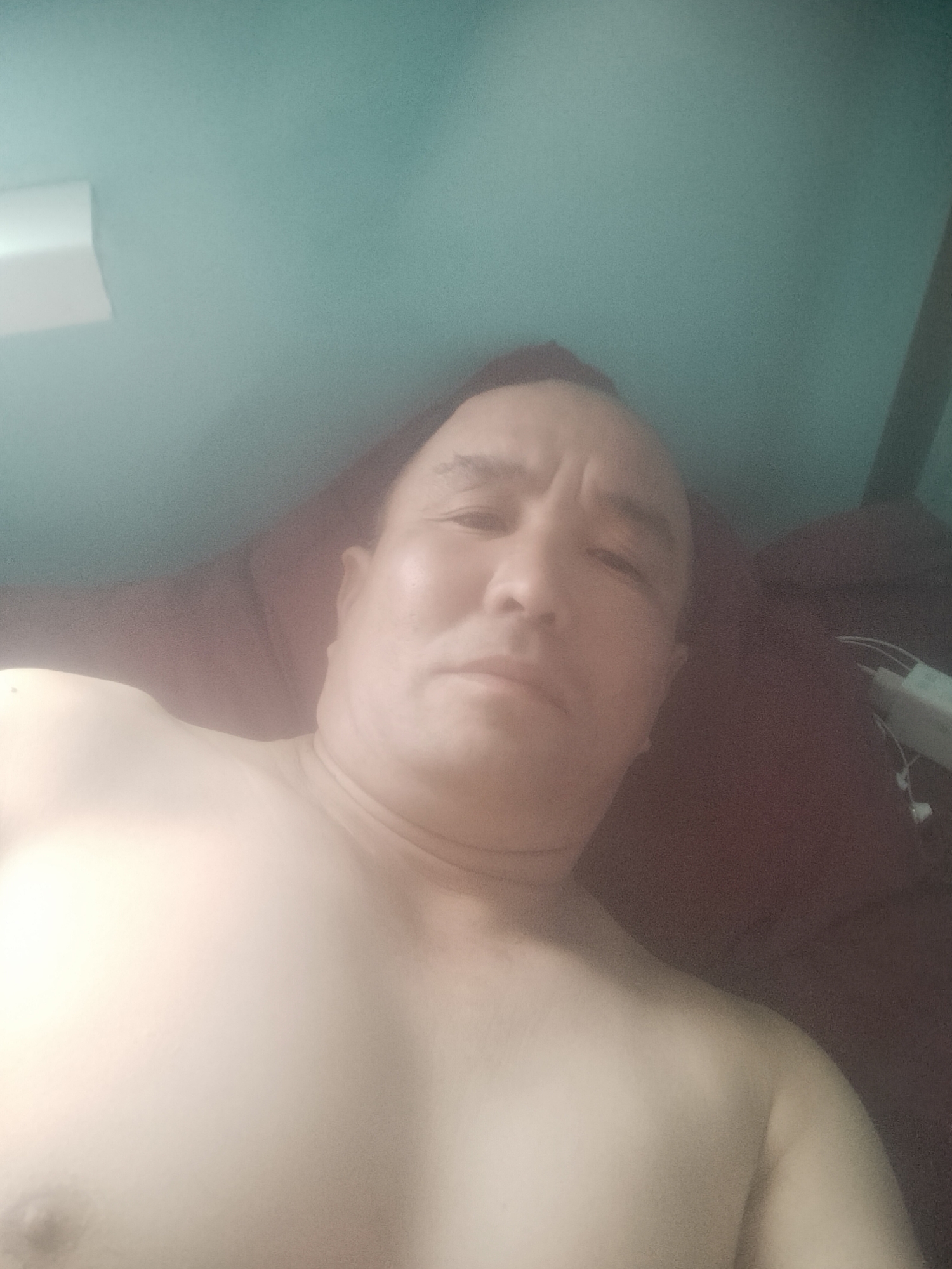 A man lies on his back, facing upwards, with his upper body visible. He has short, dark hair and appears to be of Asian descent. His eyes are looking towards the camera, and his expression is neutral. The man is shirtless, revealing his chest and shoulders. He is lying on a dark red surface, possibly a bed or sofa, and the background consists of a teal-colored wall and ceiling. A white object, possibly a light fixture or air vent, is visible on the ceiling. A dark vertical line, potentially a doorframe or furniture, is present on the right side of the image. No specific geographical markers are visible to determine the location within Jezqazğan, Kazakhstan. The lighting is dim, suggesting an indoor environment.