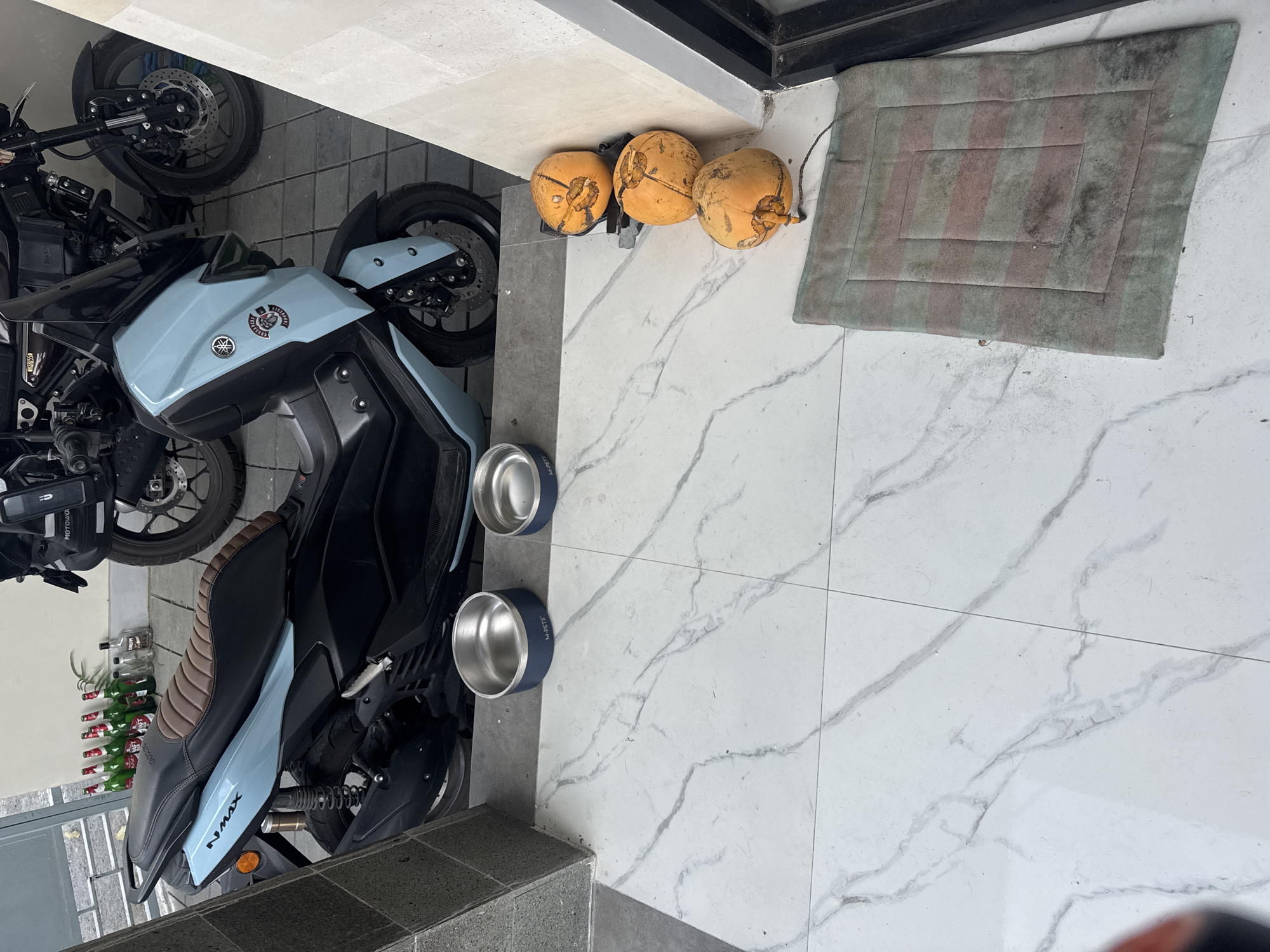 The image depicts an outdoor patio area, possibly a driveway or entryway, in Sukawati, Indonesia. The ground is tiled with a white marble-patterned material. In the background, two motorcycles are parked: a light blue Yamaha NMAX scooter with a brown seat and a black motorcycle. In front of the scooter, on the tiled floor, are two empty silver pet bowls. To the right of the bowls, against a light-colored wall, are three yellow coconuts stacked vertically. A small, rectangular mat with green and red stripes lies on the floor near the coconuts. A row of green bottles and two clear bottles are visible on a raised platform behind the scooter. The overall scene suggests a domestic or commercial setting in a tropical climate.
