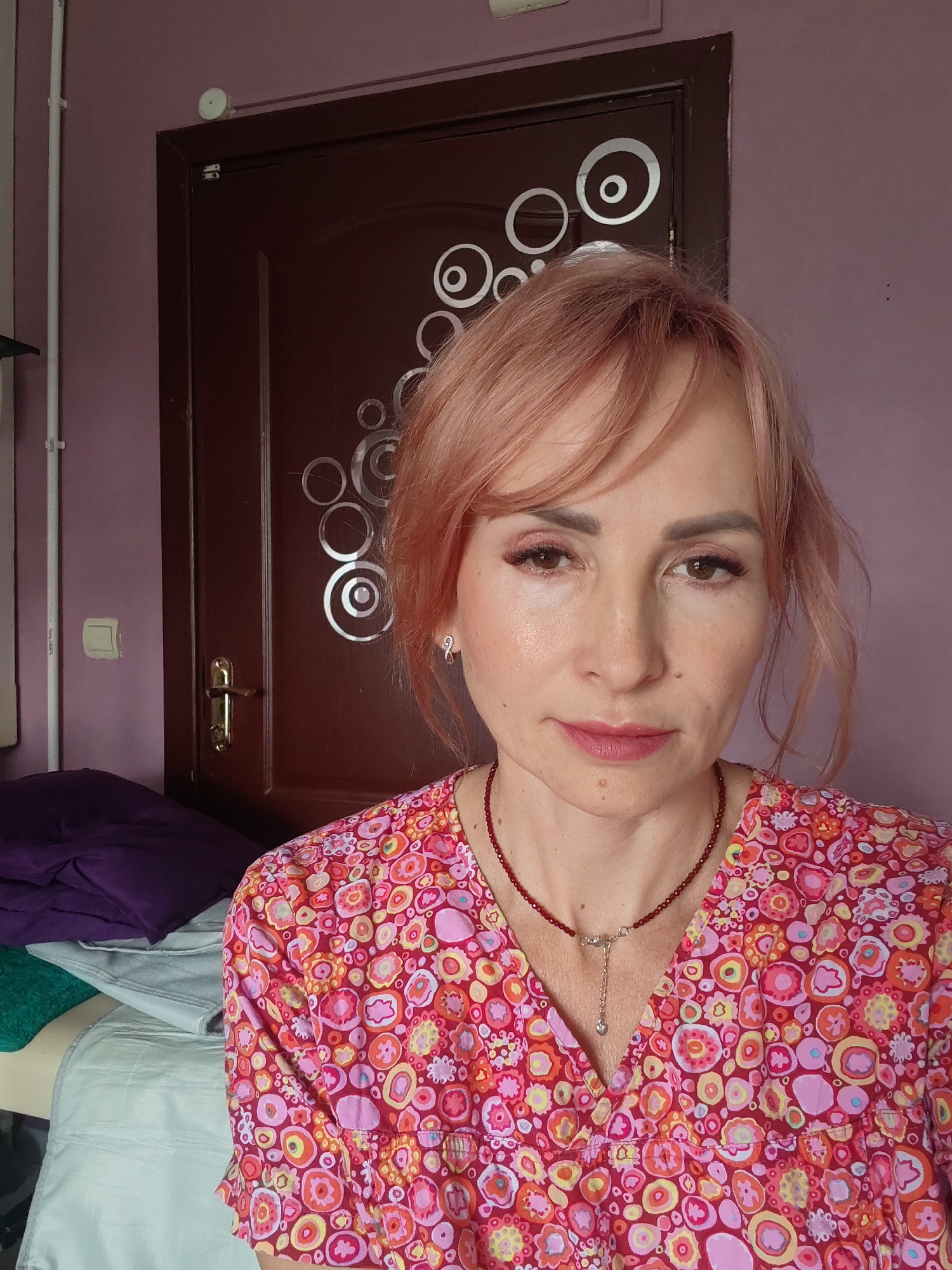 A medium shot shows a woman with short, pink-toned hair, a fair complexion, and light makeup, including eye shadow and lip gloss. She is looking directly at the camera. She is wearing a v-neck top with a colorful, abstract floral print in shades of pink, orange, yellow, and red. A thin, dark red beaded necklace with a silver pendant is visible around her neck.
The woman is positioned in what appears to be a room. Behind her, a dark brown door with a brass handle is visible. The door has a decorative pattern of concentric circles in a silver, reflective material. To the left of the door and behind the woman, a section of a wall painted in a light purple hue is visible. Mounted on the wall is a light switch. In the lower left corner of the frame, partially visible, are stacked items that appear to be folded textiles in shades of purple, teal, and light blue, resting on a surface. The overall lighting suggests an indoor setting, possibly a domestic or treatment room environment. No specific landmarks of Chaikovsky, Russia, are identifiable in the image.