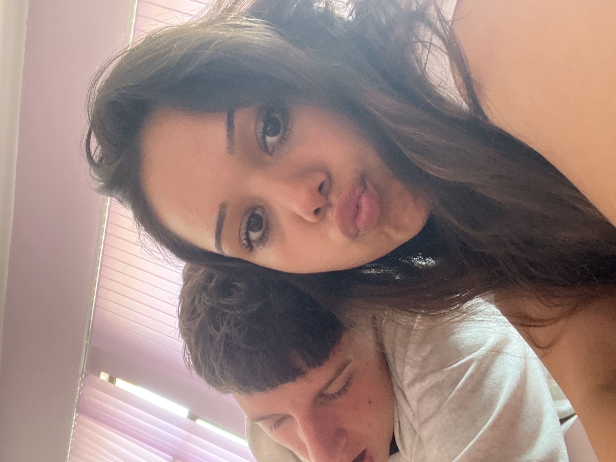 The image is a high-angle, close-up selfie featuring two individuals in an indoor setting. The scene is dominated by the faces and upper bodies of the subjects, with a soft, pale background.
The person positioned at the top of the frame, appearing to be female, looks directly at the camera with a playful expression, puckering her lips. She has long, dark brown hair that falls around her face and over the second person. Her dark eyes are open and focused, and she has neatly shaped eyebrows. Her left shoulder and arm are visible on the right side of the image.
Beneath her, and partially obscured by her hair, is a second person, appearing to be male. His face is turned downwards and to the left, with his eyes mostly closed or squinting, and his mouth slightly ajar. He has short, dark brown hair with bangs across his forehead. He is wearing a light-colored, possibly grey or white, t-shirt or sweatshirt. His relaxed posture suggests he might be resting or sleeping.
The background shows a pale lavender or pink-tinted wall and Venetian blinds, indicating an indoor environment, likely during daytime given the ambient light. The overall mood is intimate and casual. No text is visible in the image.