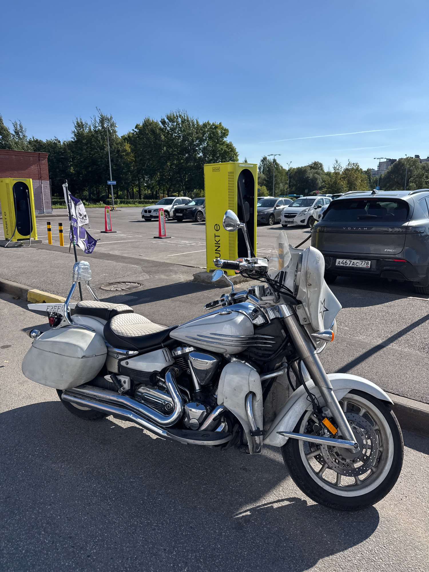 A white cruiser motorcycle with saddlebags and flags is parked in a lot with electric vehicle charging stations in Vsevolozhsk, Russia. The motorcycle is in the foreground, partially obscuring a dark grey Voyah electric SUV, which is plugged into a yellow charging station. Several other cars and charging stations are visible in the background, along with trees and buildings. The sky is clear and blue.