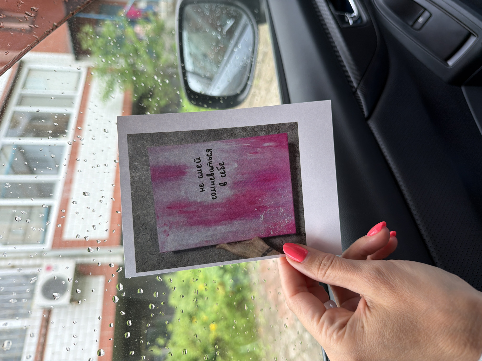 The image captures a close-up scene from inside a vehicle, looking out through a rain-splattered window. A person's hand, with bright pink/coral nail polish, holds up a white rectangular card.
The card displays a smaller, framed image featuring an abstract background of pink and magenta hues with some white streaking. Written in black text on this background is the Russian phrase: "не смей сомневаться в себе", which translates to "Don't dare doubt yourself."
Through the car window, an exterior view is visible. On the left, a building with white-framed windows and orange-toned architectural trim is partially seen, along with an outdoor air conditioning unit. Green foliage, likely from trees or bushes, is blurred in the background, all viewed through the water droplets on the glass, indicating rainy weather. On the right, the dark interior of the car is visible, including a textured door panel and a silver door handle, along with a blurred reflection in the side mirror, which also has raindrops on it. The scene appears to be set during the daytime in Birobidzhan, Russia, suggesting a moment of personal reflection or encouragement within a car.