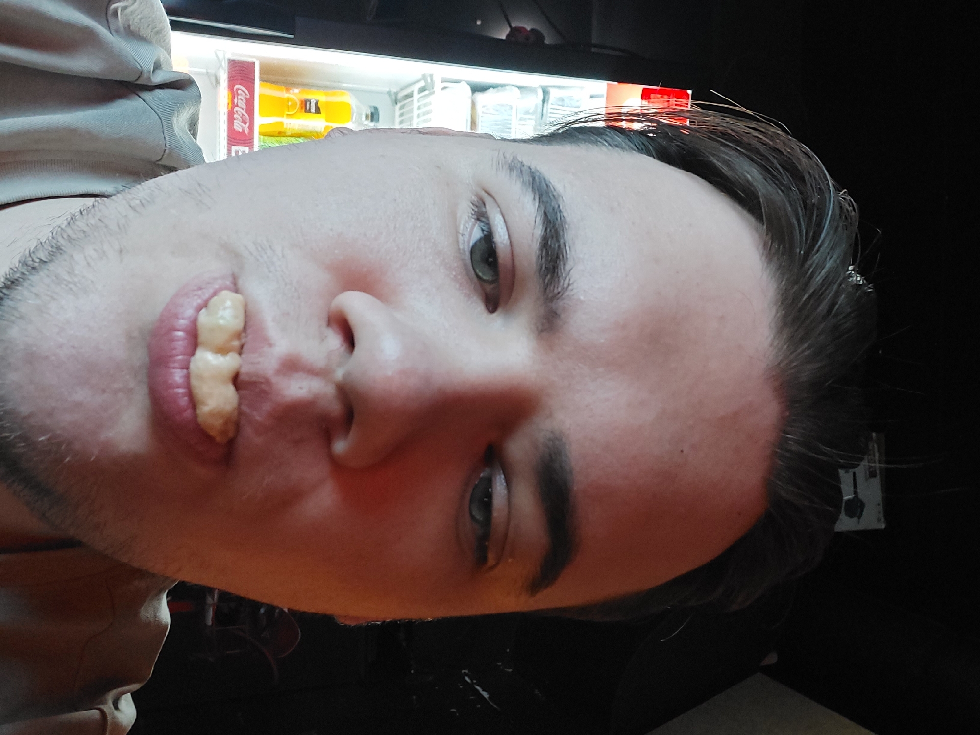 A man with light skin, short brown hair, and stubble is holding a piece of food in his mouth and looking at the camera. He is positioned in front of a vending machine that contains beverages. The location is not discernible as Tashkent, Uzbekistan based on the image's content alone.