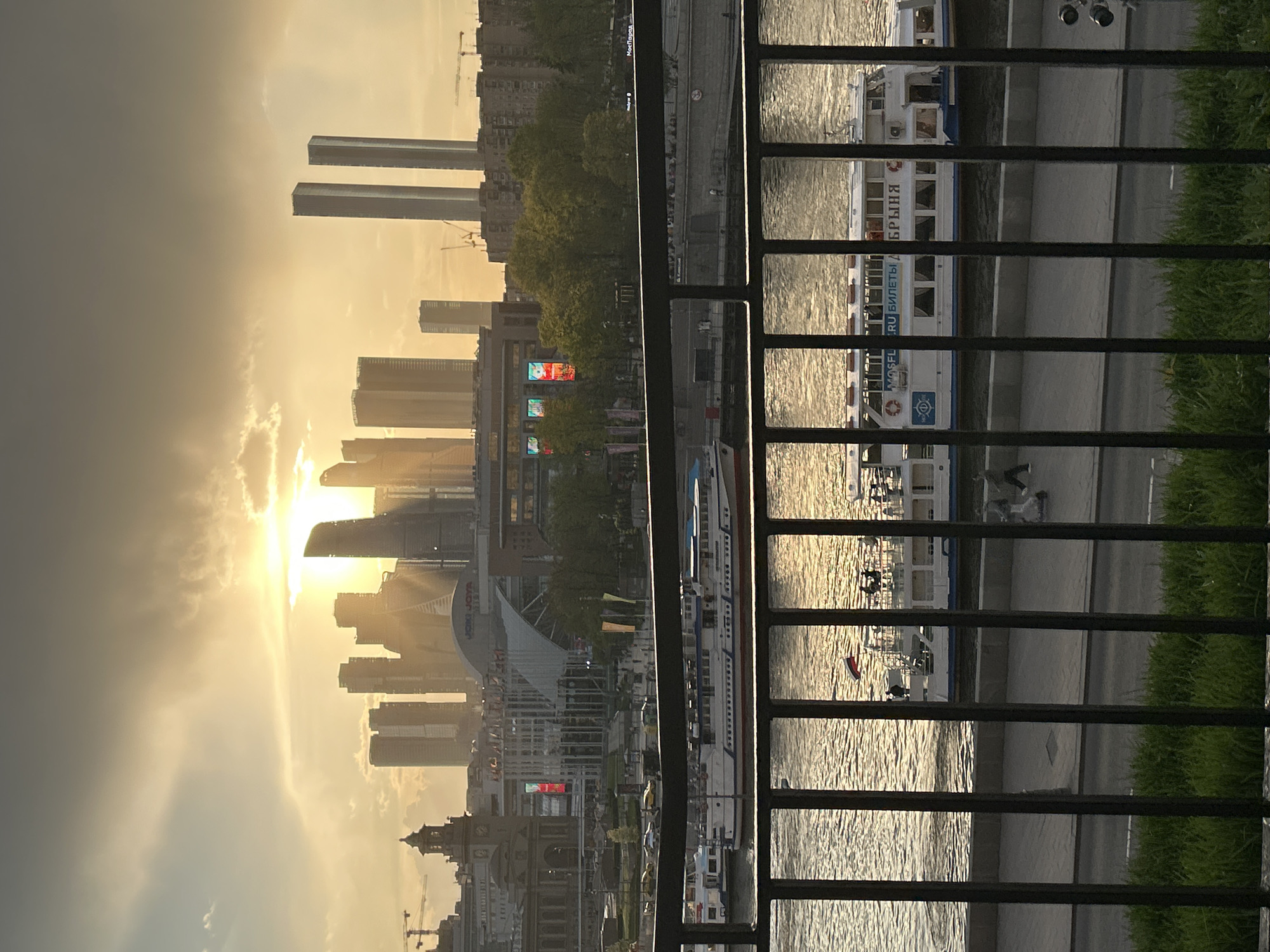 The image shows a cityscape at sunset, viewed from behind a dark, metal railing. In the background, buildings of various heights are silhouetted against the setting sun. The sun is shining brightly, casting rays over the buildings and the sky.
A body of water, potentially a river, is visible in the middle of the image. Several boats, predominantly white with red accents, are docked along the riverbank.
The scene is potentially located in Moscow, Russia. This is based on the buildings, river, and style of the boats. Also, boats are shown to have the name “MOSFLOT” on their side.