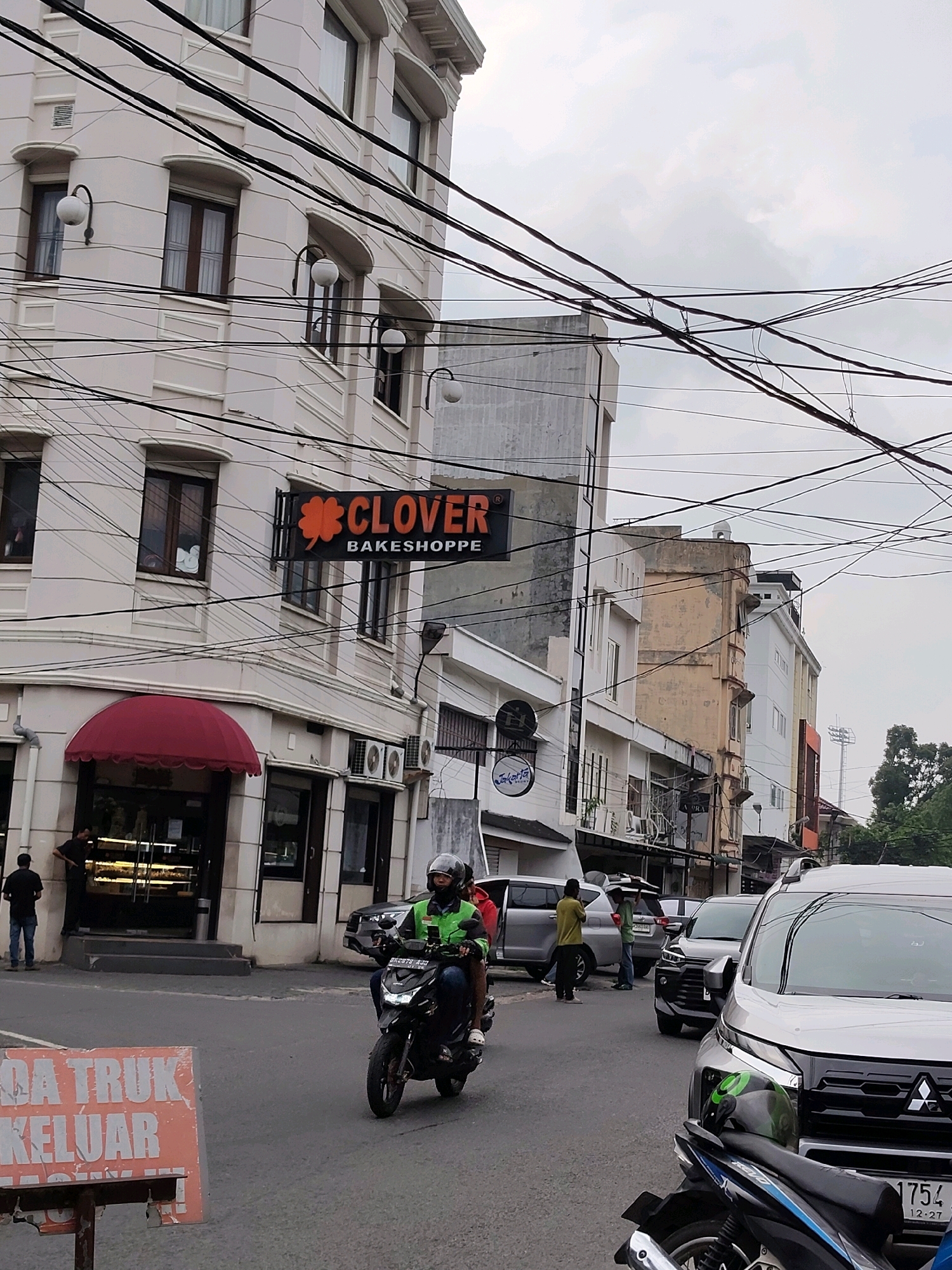The image depicts a street scene in the city of Medan, Indonesia. In the foreground, a person is riding a motorcycle, wearing a green jacket and helmet. To the left, a sign with orange text on a white background reads "DA TRUK KELUAR". Behind the motorcycle, several cars are parked or driving, including a silver SUV in the foreground on the right.
Dominating the left side of the image is a multi-story building with a light-colored facade. A sign reading "CLOVER BAKESHOPPE" with an orange clover logo is visible on the building. Below the sign, a shop with a red awning and glass doors is present, with a person standing near the entrance.
Other buildings of varying heights and architectural styles line the street, some appearing older with weathered facades. The sky is overcast. Numerous electrical wires crisscross overhead, creating a dense network. The environment suggests an urban setting with commercial activity.