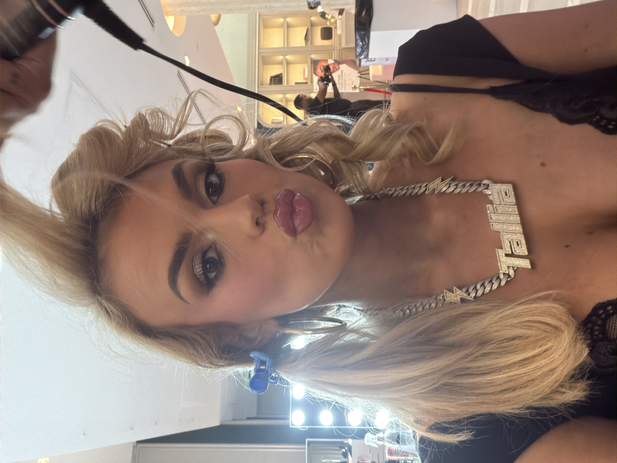 A blonde woman with a full face of makeup puckers her lips for a selfie. Her hair is styled with curls and a blue clip. She wears a silver necklace. The background includes a makeup vanity with lights, and a retail area with shelves. A man is seen holding a camera, potentially a photographer, in the background. The location is likely Miami Beach, United States of America.