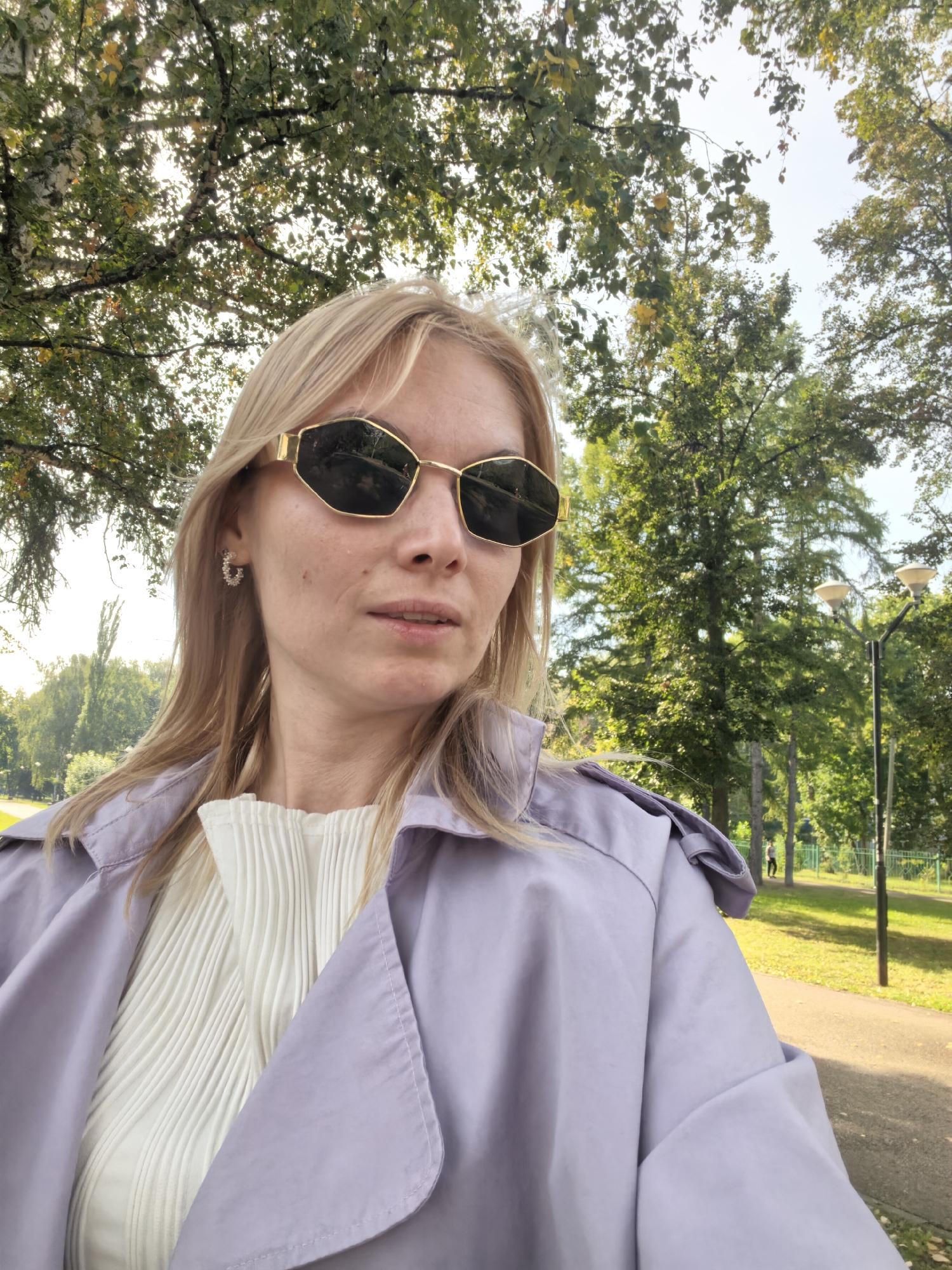 A blonde woman wearing a light purple trench coat over a white pleated top and geometric sunglasses is taking a selfie outdoors. She has light makeup and is wearing small hoop earrings. The background is a park with lush green trees and a pathway. In the background, there are two lampposts and a person walking on the path. The sun is shining brightly, casting shadows. This scene appears to be set in the city of Sterlitamak, Russia.