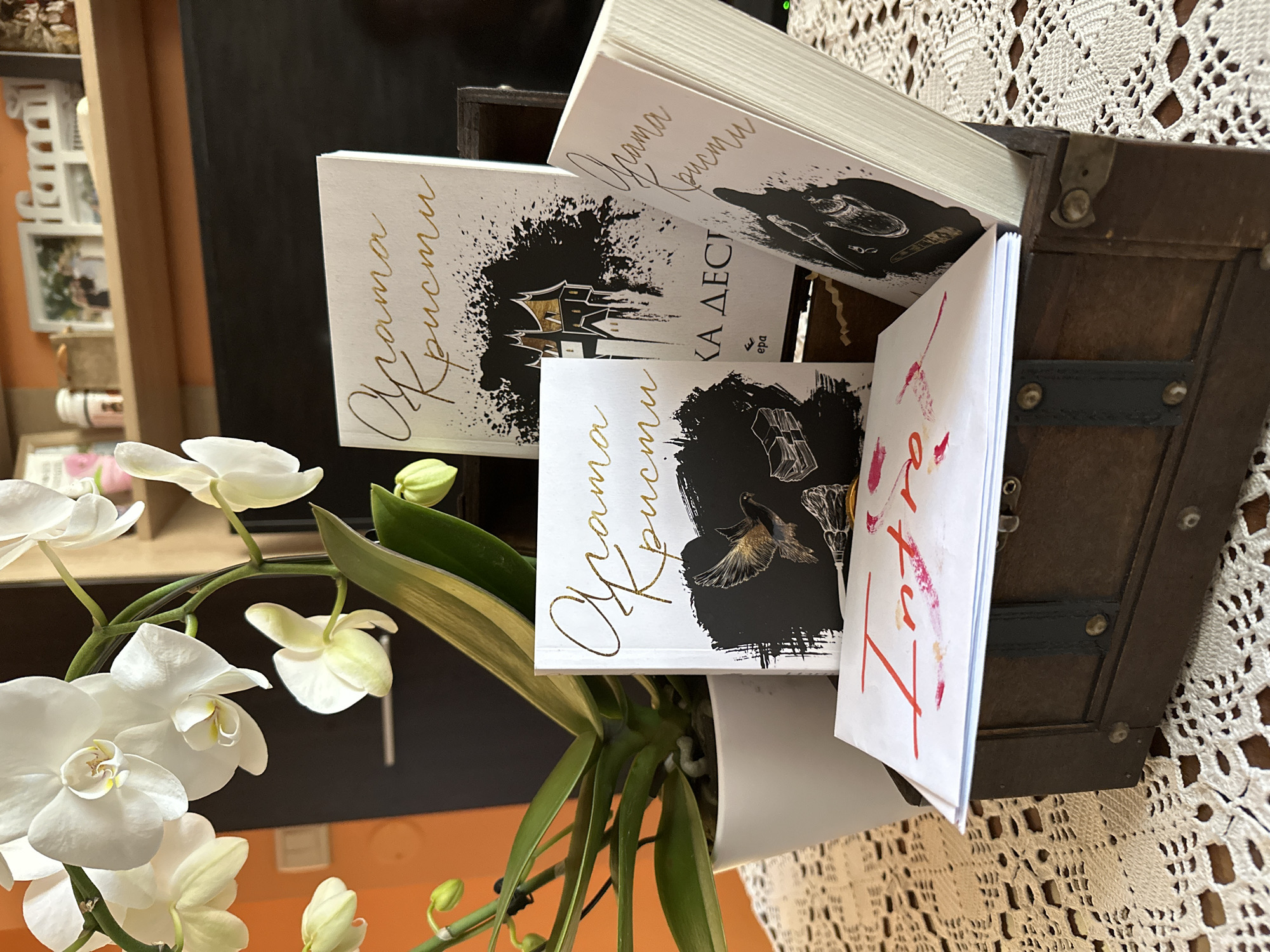 The image shows a collection of books with the text "Qrama Khristu" on the cover, sticking out of a small wooden chest. The books have different cover illustrations in black ink. To the left of the chest, there's a white orchid plant in a white pot. The chest and orchid sit on a table covered with a white lace tablecloth. In the background, a partially visible wooden shelf unit and a black object are visible. The setting appears to be indoors, potentially a home. It is not possible to verify that the image was taken in Sofia, Bulgaria, based on the provided information.