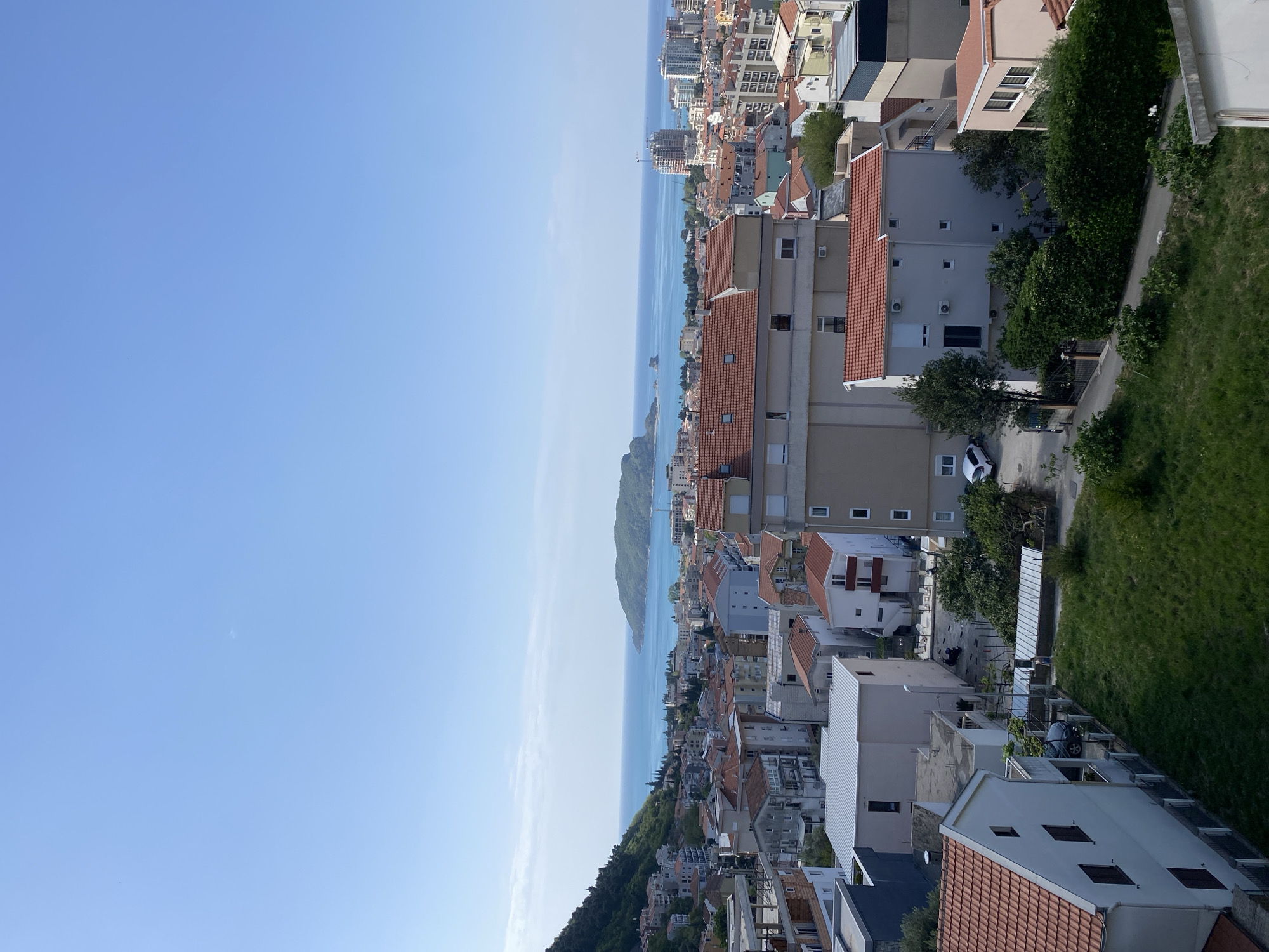The image displays a panoramic view of a coastal town, likely Budva, Montenegro, spread across a hillside overlooking a serene blue sea. The scene is captured on a bright, clear day, suggesting mid-morning or early afternoon, with abundant sunlight illuminating the landscape.
In the foreground and midground, a dense cluster of buildings with distinct red and orange tiled roofs dominates, characteristic of Mediterranean architecture. These structures vary in size and style, ranging from multi-story residential buildings to what appear to be commercial establishments. Some buildings have light-colored facades, and air conditioning units are visible on their exteriors. Roads and pathways wind through the urban fabric, and a few white cars can be seen parked or driving on them. Green trees and lush bushes are interspersed among the buildings and along the natural slopes, adding pockets of vegetation to the built environment.
The expansive Adriatic Sea occupies the midground, its calm, blue waters stretching towards the horizon. A distant landmass, possibly an island or peninsula, is visible on the far side of the bay, contributing to the picturesque backdrop. The sky above is a clear, uninterrupted expanse of light blue, devoid of clouds, indicating excellent weather conditions. To the far right of the frame, a steep, vibrant green grassy slope descends, outlining the town's hilly terrain. There are no people visible, and no specific activity or event is taking place, presenting a tranquil, static landscape. No visible text is discernible in the image.
