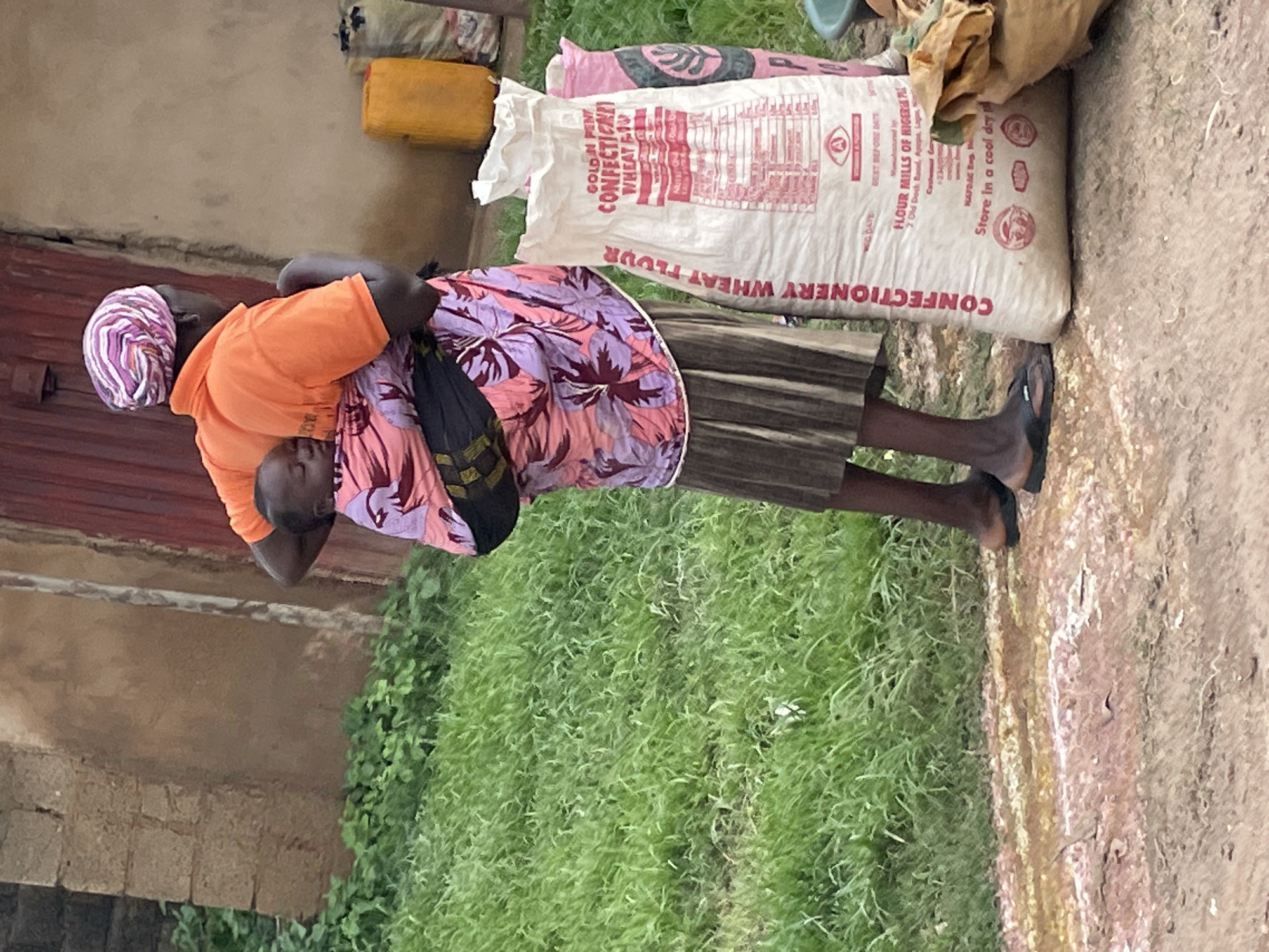 A Black woman wearing an orange short-sleeved shirt and a pink and purple headscarf carries a baby on her back. The baby is wrapped in a pink and purple patterned fabric and is asleep. The woman is wearing a brown pleated skirt and black sandals. She is standing on a dirt path next to a grassy area. To her right are several large sacks of flour, one of which is white with red lettering. Behind her is a building with a red door and a tan wall. There is a yellow plastic container to the right of the flour sacks, and other sacks are visible behind it. The scene appears to be in Wang, Nigeria.