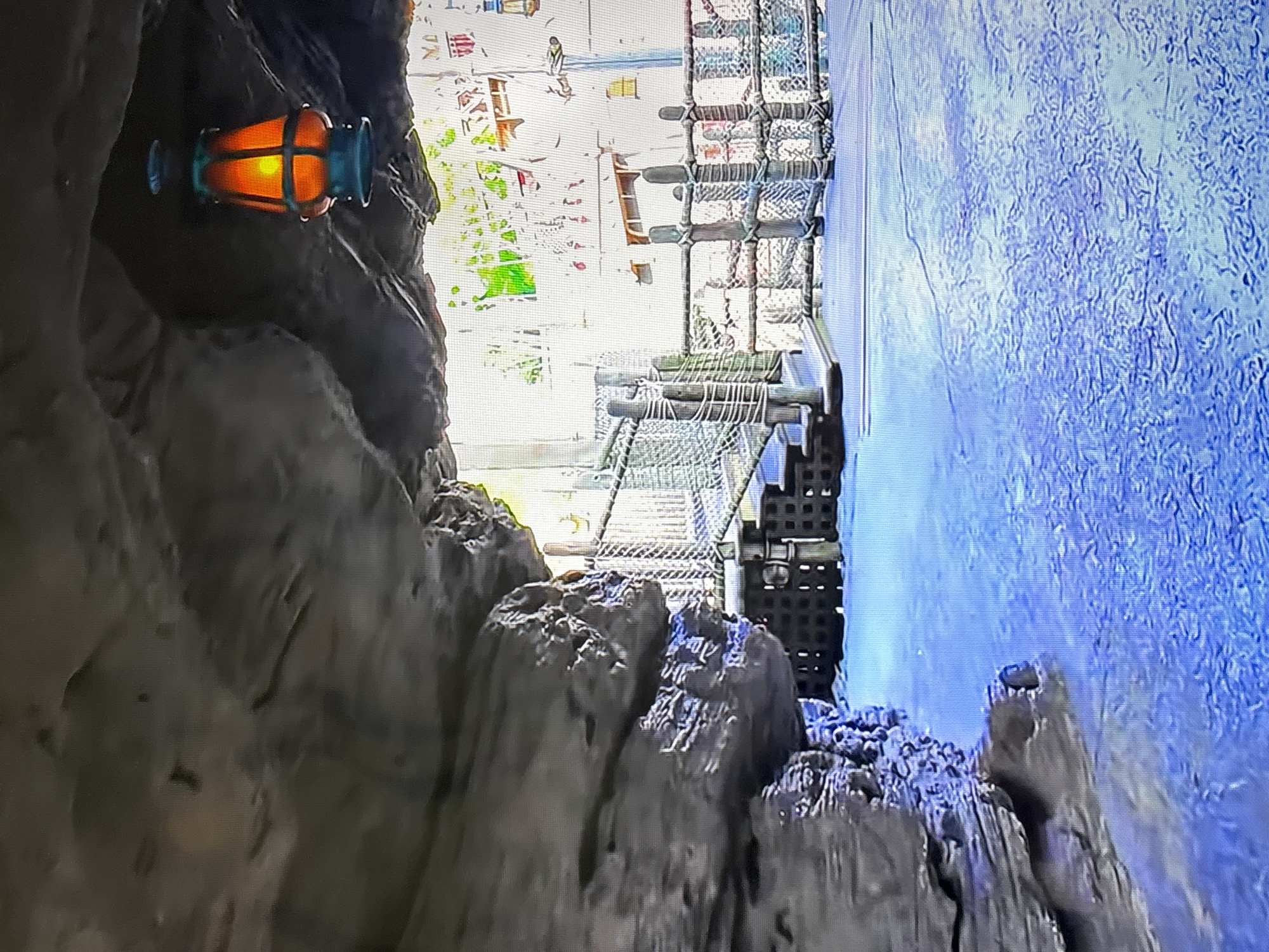 The image depicts a view from a cave-like structure opening onto a scene of a cityscape and water. A lantern is affixed to the cave wall. Outside the cave, a rope bridge or walkway is visible. Beyond that, there is a cityscape, partially obscured by bright light, possibly sunlight. To the right of the bridge is a body of blue, textured water. The location and city are not identifiable.