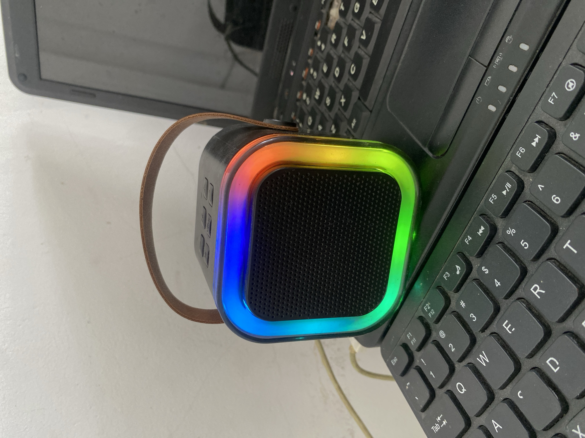 A small, black, square speaker with a brown leather handle sits on top of a black laptop keyboard. The speaker has a colorful LED light that transitions through blue, green, and red along its edges. The laptop is open, and its screen is visible in the background. The scene appears to be indoors, possibly on a desk, with a white wall in the background. The image is taken from a slightly elevated angle.