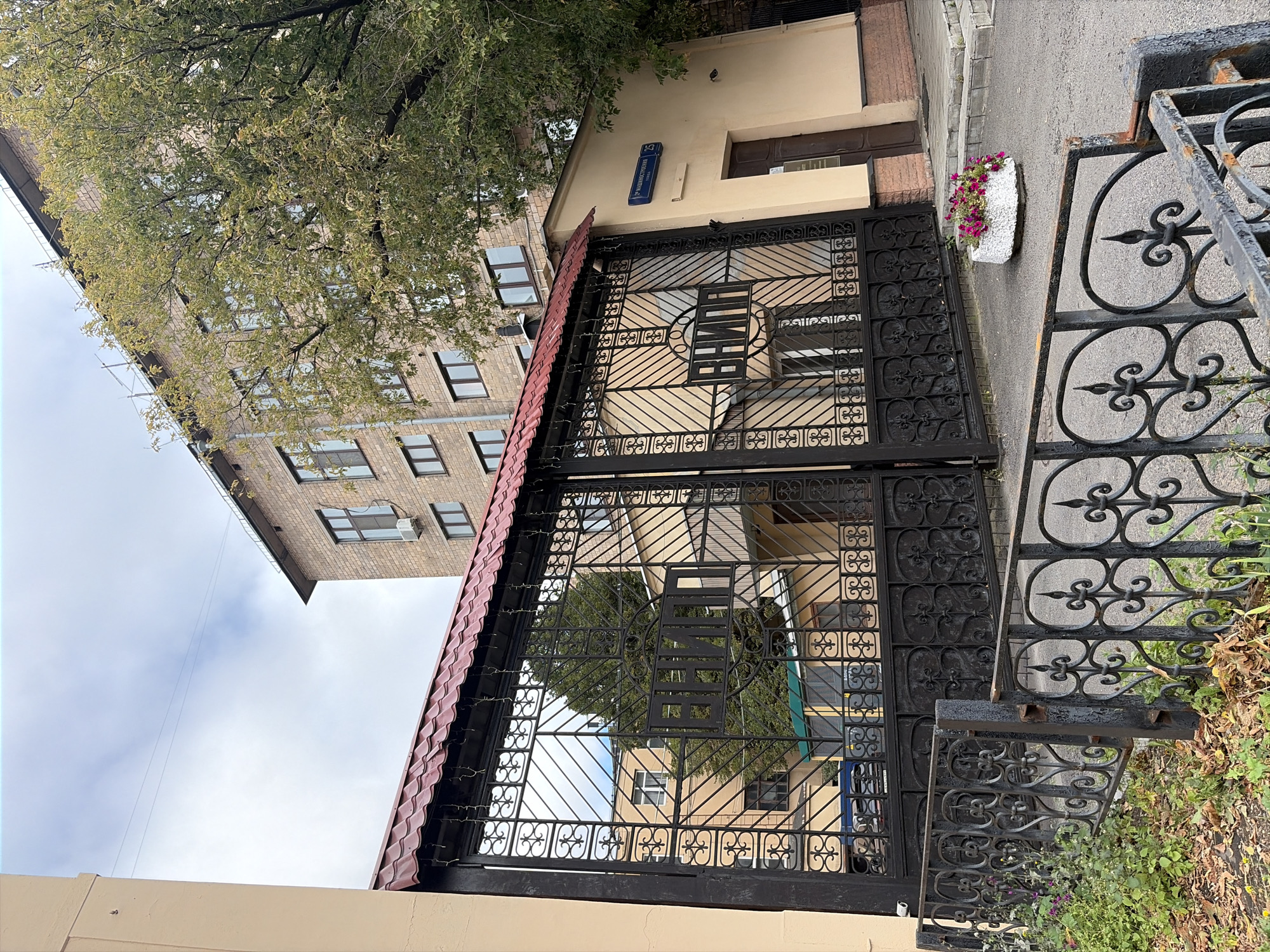 The image shows the entrance gate to a building complex, likely an institute or research center, in Moscow, Russia. The gate is ornate, made of black wrought iron with a red tiled roof overhead. The letters "ВНИИ" are prominently displayed in the center of the gate. Behind the gate, several buildings are visible, one made of light-colored brick and another with a yellow facade. A street sign indicating "21-я Магистральная улица, 25" is affixed to the yellow building, identifying the location. A planter with pink and purple flowers sits on the paved ground in front of the entrance. The sky is overcast.