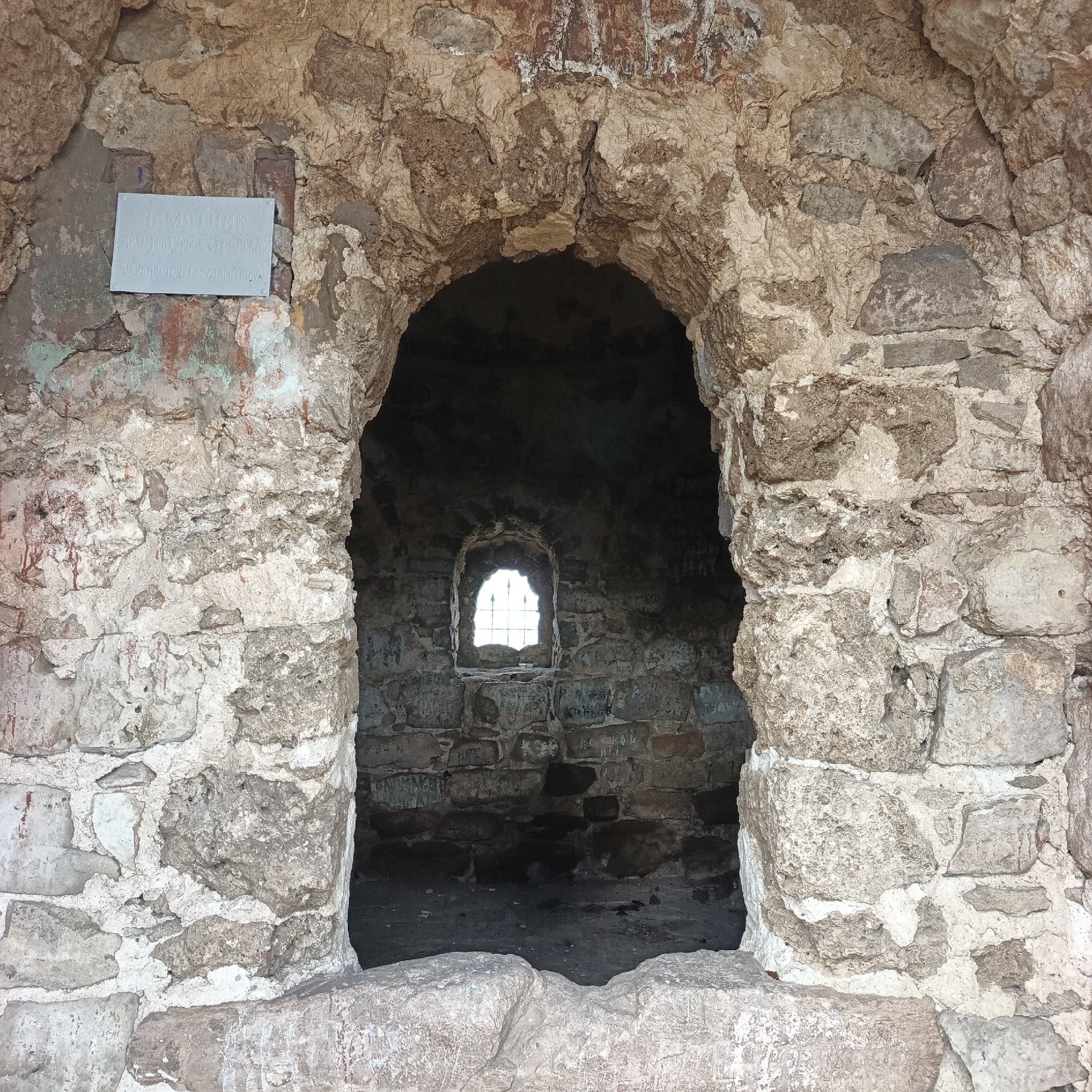 An external frontal medium shot captures the entrance to a stone structure located in Verkhnie Termy, Russia. The entrance is an arched opening framed by rough-hewn stones, some of which are weathered and appear to have remnants of faded paint or plaster. The stone masonry is irregular and shows signs of age and erosion.
To the left of the entrance, mounted on the stone wall, is a light-colored plaque with inscribed text. The text is partially obscured by the stone texture and angle, but it appears to be in Cyrillic script and reads: "ПАМЯТНИК АРХИТЕКТУРЫ XII-XX BEKA, ОХРАНЯЕТСЯ ГОСУДАРСТВОМ" which translates to "ARCHITECTURAL MONUMENT OF THE 12th-20th CENTURY, PROTECTED BY THE STATE."
The opening leads to a darker interior. Visible through the entrance is a small, arched window in the back wall of the structure. This window has a metal grate or bars across it and allows some light to penetrate the interior, illuminating the rough stone construction of the back wall and the floor. The floor of the interior appears to be dirt or rough stone. Some graffiti or inscriptions are visible on the interior stone walls. The foreground consists of large, rough stones forming a kind of threshold or base for the structure. The overall impression is one of an ancient, weathered historical site.