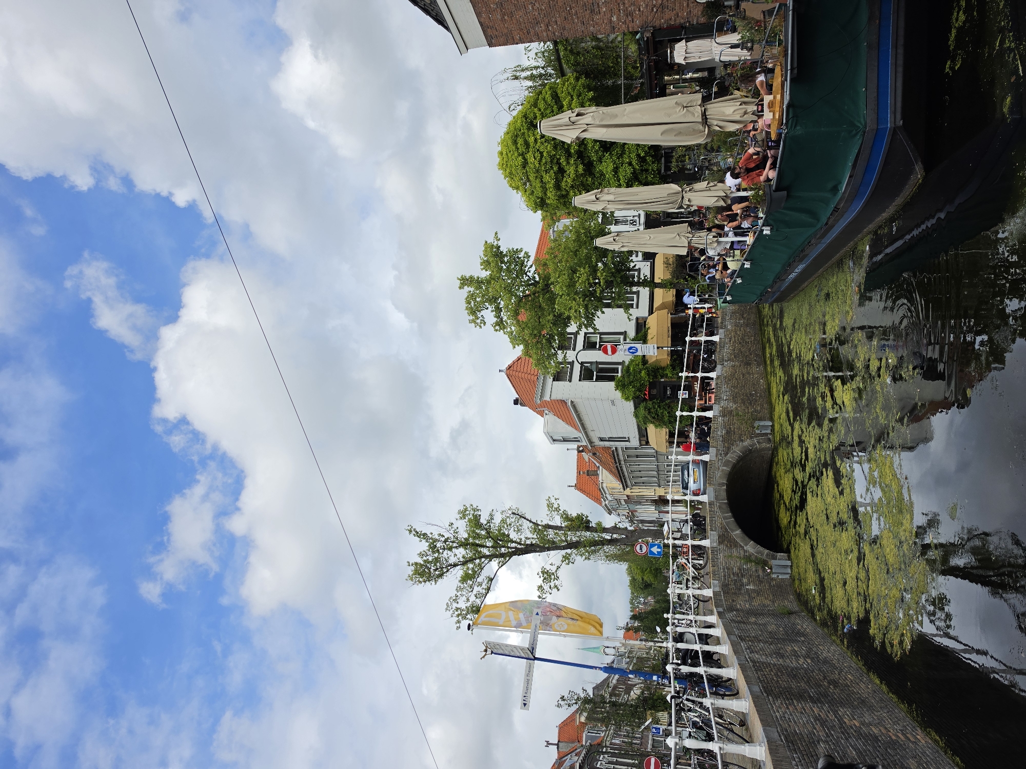 The image depicts a picturesque canal scene in Delft, Netherlands, on a cloudy day. A bridge with an arched opening spans the canal, with a brick retaining wall and a white railing along its edge. Several bicycles are parked along the railing.
In the canal, a green-covered boat is docked, occupied by a group of people. The boat appears to be a floating cafe or restaurant, with tables and chairs visible on its deck, some of which are shaded by large, closed umbrellas. The canal water is murky and covered with a layer of green algae.
The background features buildings with terracotta roofs, characteristic of Dutch architecture, and lush green trees. A tall tree stands prominently near the bridge, and a yellow flag with an image is visible on a pole. Street signs and a traffic light are also present, indicating a public street along the canal. The overall impression is a vibrant urban landscape blending historical architecture with modern leisure activities.