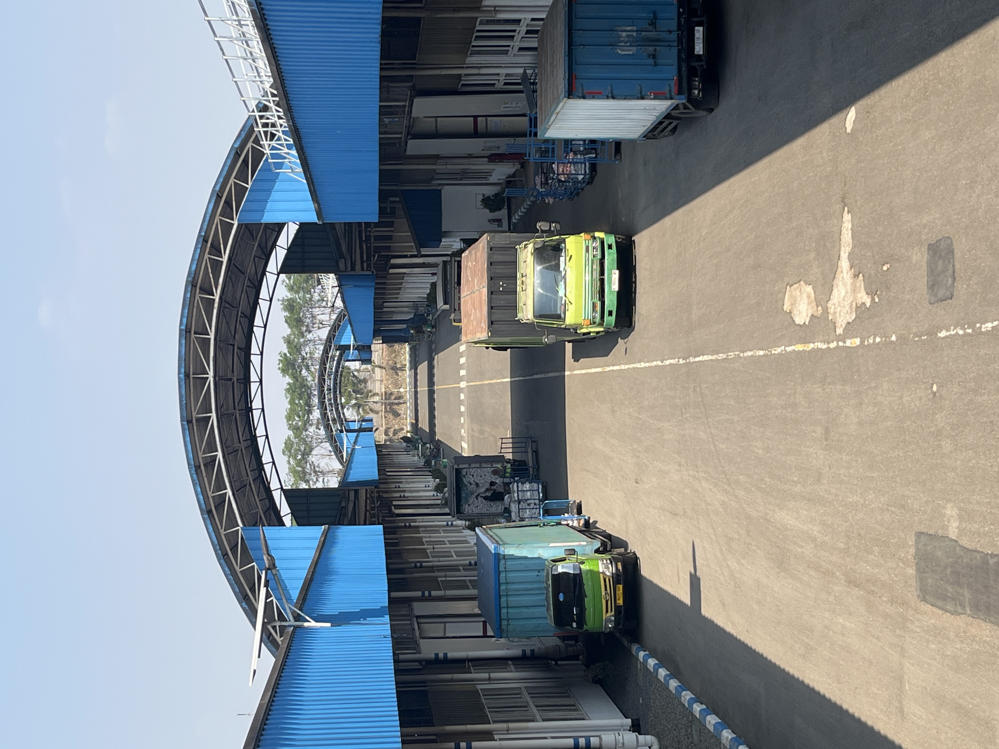 The image depicts an outdoor loading dock area with several trucks and cargo. The scene is located in Pelang, Indonesia. The architecture features long, blue-sided buildings with a high, arched, open-air canopy extending over the central road. The sky is clear and blue, indicating daylight.
On the left side of the road, a light green truck with a blue cargo container is parked near one of the buildings. Further down, two more trucks are visible, one with a dark gray cargo container and another with a light gray, open-top container. In the foreground on the right, a blue truck with a white cargo container is parked.
Several individuals, likely workers, are visible near the trucks. Some appear to be loading or unloading cargo, with trolleys and boxes present. The road is paved and marked with white lines. Shadows from the buildings and canopy are cast across the pavement. The overall impression is of a functional logistics or distribution center.