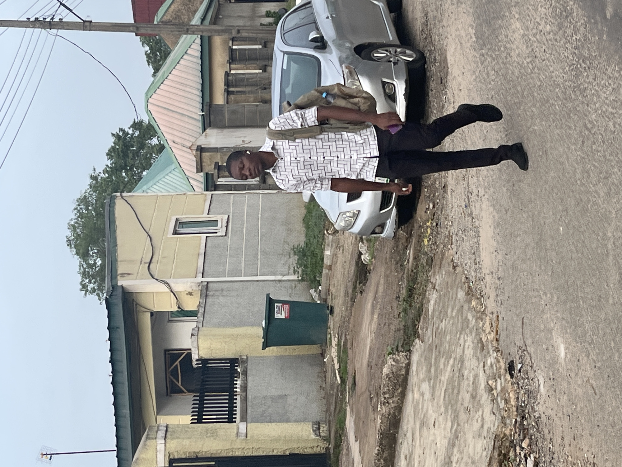 This media file captures an outdoor street scene in what appears to be an urban or suburban setting in Abuja, Nigeria. The primary subject is a young man, likely in his 20s or 30s, with dark skin, standing on a paved road. He is wearing a short-sleeved white and black patterned shirt, dark trousers, and dark shoes. A light-colored backpack or bag is slung over his left shoulder, and he holds a purple object, possibly a phone, in his left hand. An earbud appears to be in his right ear. He looks directly towards the viewer with a neutral expression, his right arm slightly bent.
Behind the man, a silver sedan car is parked on the side of the road. To the left and right, several residential buildings with light-colored walls (yellow and gray) and varying roof types (some with corrugated metal, others flat) line the street. Overhead power lines are visible against a light, overcast sky in the upper left of the frame. Further down the street, near one of the buildings, a dark green cylindrical trash can is visible. On this trash can, a red and white label with partial text reads "LAGOS STATE...". The road surface is paved but appears somewhat uneven, with a dirt and gravel shoulder. Sparse trees and greenery are seen in the background behind the buildings. The lighting suggests it is daytime, possibly a cloudy day or early morning/late afternoon.