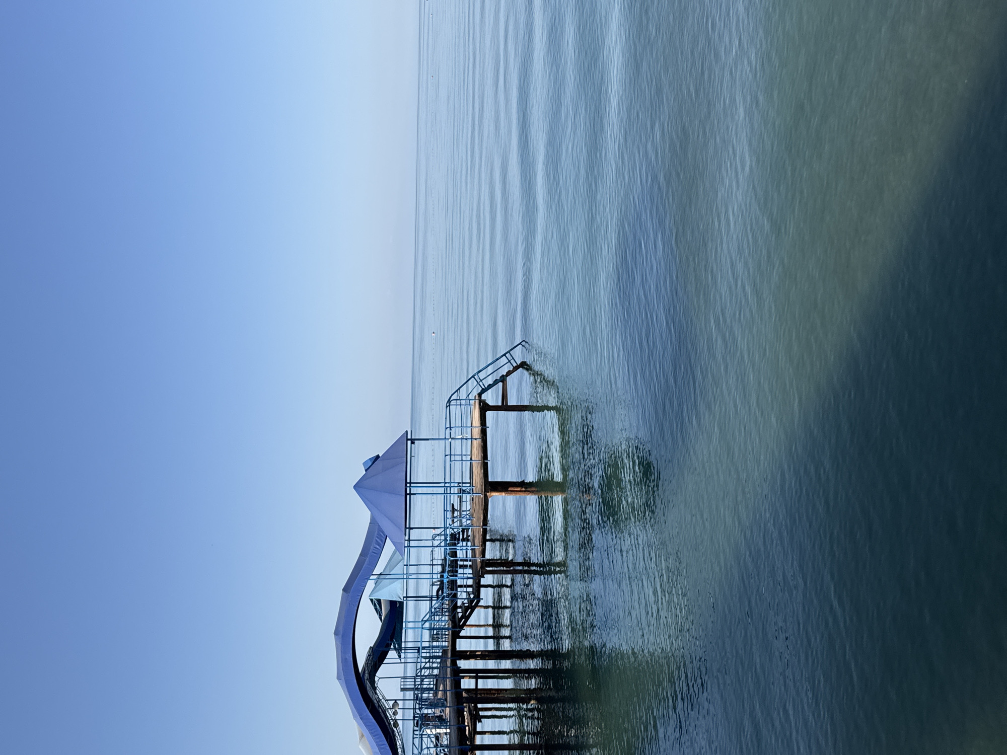 An elevated view captures a wooden pier with blue canopies extending over a calm, blue body of water. The pier is supported by thick, brown posts submerged in the water, and features railings and stairs leading down to the water's edge. The sky above is a clear, bright blue, with the horizon line a faint blur in the distance. The water reflects the pier and the sky, creating a sense of depth and tranquility. The location is the city of Sary-Oy, Kyrgyzstan.