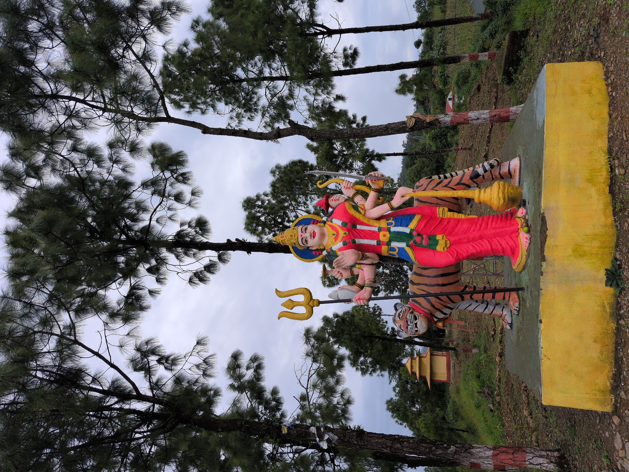 In Kapurkot, Nepal, a vibrant statue of a multi-armed goddess, possibly Durga, stands prominently. The goddess is depicted with at least ten arms, holding various symbolic objects including a trident, a sword, and a mace. She is adorned in a red sari with blue and green accents, a golden headdress, and intricate jewelry. The statue is positioned atop a tiger, a common mount for Hindu deities, which is rendered in bold orange and black stripes. The entire sculpture rests on a bright yellow platform, which in turn is situated on a concrete base. The background features a dense arrangement of tall pine trees with sparse foliage against a cloudy sky. In the distance, a small, tiered yellow and red temple structure is visible, surrounded by lush green hills and vegetation. The ground around the statue is uneven, covered with dirt, rocks, and patches of grass. The trees in the foreground have their lower trunks painted with red and white bands.