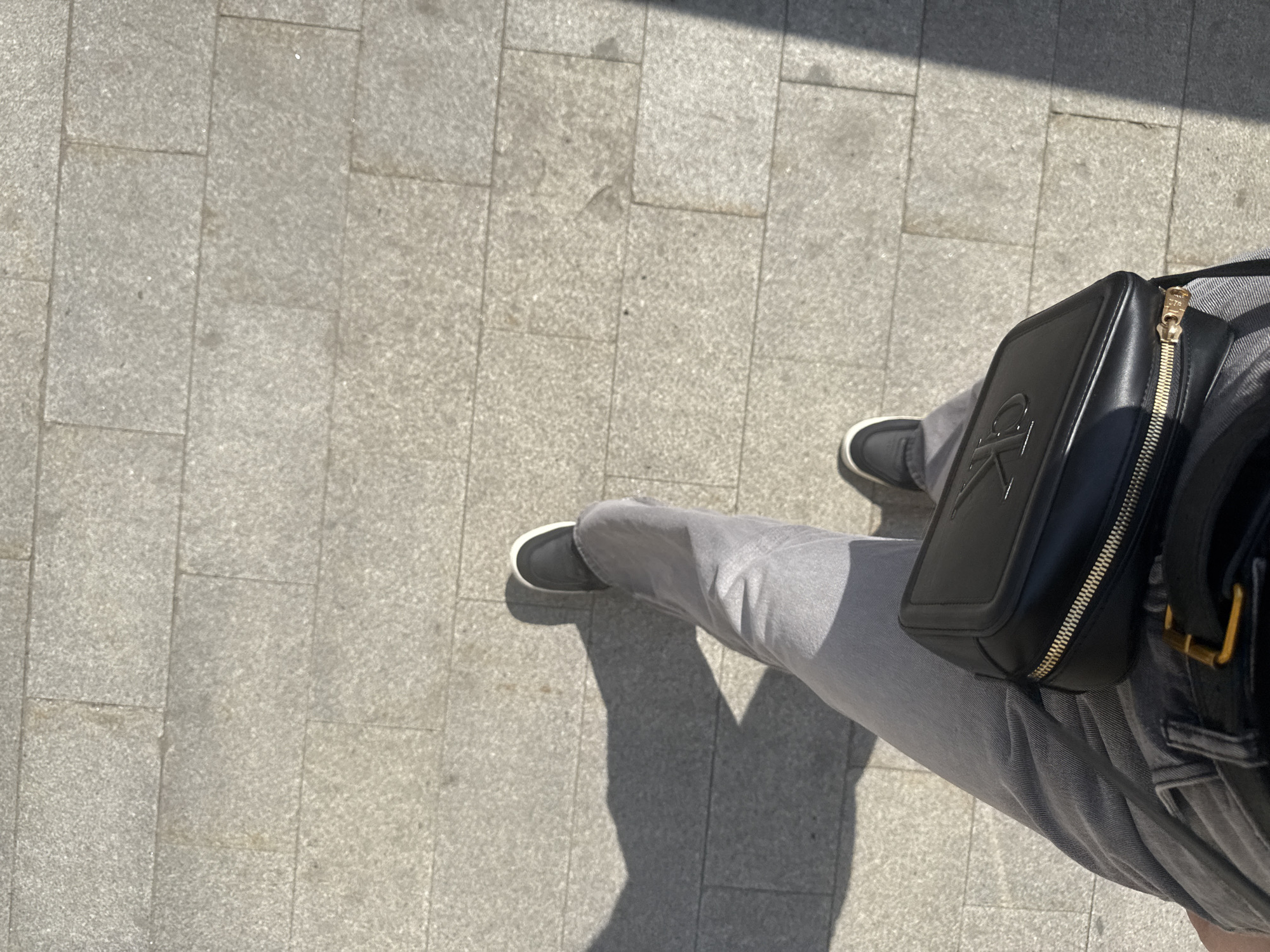 The image is a top-down shot of a person standing on a paved surface, likely a sidewalk or pedestrian area. The paving is made of light gray rectangular stones. The person is wearing gray jeans and dark-colored shoes with white soles. A black leather crossbody bag with a gold zipper and the letters "CK" embossed on the front is visible. The scene suggests an urban setting, possibly in Moscow, Russia.