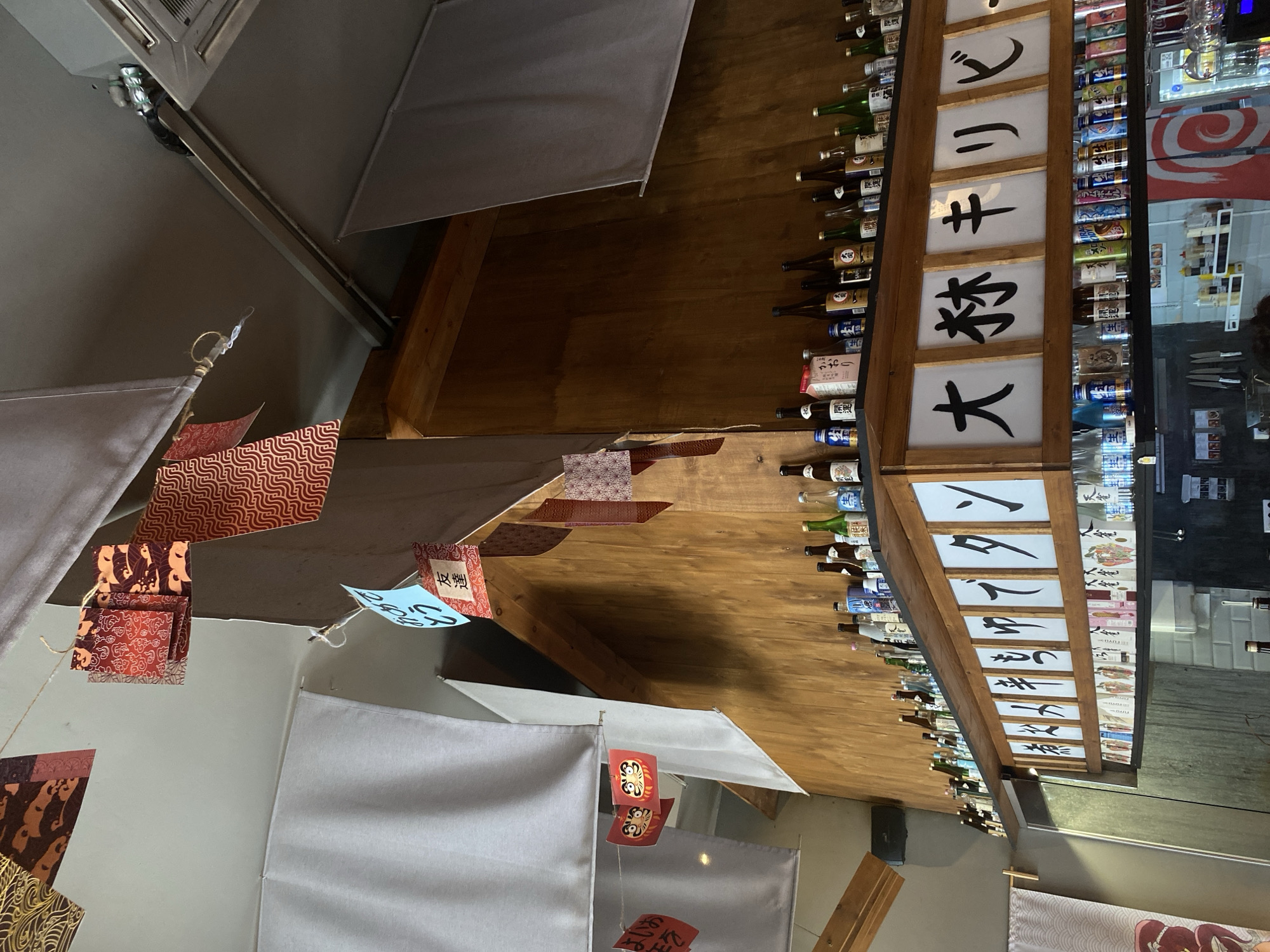 This indoor shot reveals elements of Japanese-themed decor. The focal point is a display of sake bottles positioned adjacent to vertical wooden panels featuring Japanese script. The sake bottles are arranged in multiple rows along the length of the wooden panels. The panels have black characters that translate to: "Congratulations", "Friends", "Good", "Flavor", "Scent", "Open Luck", "Raw", "Heavenly Sparrow", "Raw", and "Ram Bottle".
Hanging below is a series of decorative banners, some white, others patterned, suspended on strings. The banners alternate with folded paper crafts in red and brown hues. There are other elements: decorative red blocks with cartoonish faces and Japanese inscriptions. The overall composition gives the impression of a bar or restaurant with a specific focus on Japanese beverages and culture. The location is not identifiable as Krasnoyarsk, Russia without additional context.