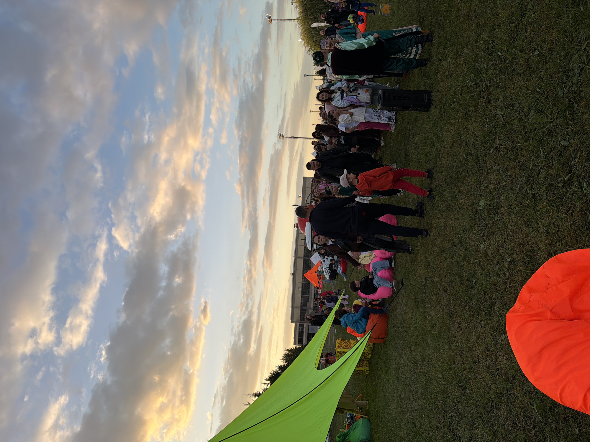 The image depicts an outdoor gathering with a crowd of people on a grassy field under a cloudy sky with hues of orange and yellow from a setting sun. A large, bright lime green canopy is set up on the left side of the frame, casting a shadow. Several beanbag chairs in various colors, including pink, orange, and green, are scattered on the grass, some occupied by people. In the background, a building with rows of windows and a large parking lot with vehicles are visible, along with lampposts and trees. The crowd appears to be a mix of adults and children, with some individuals standing and talking, while others are seated on the beanbags. A black speaker is placed on the ground in the middle of the scene. The overall atmosphere suggests a casual outdoor event, possibly a festival or community gathering.
