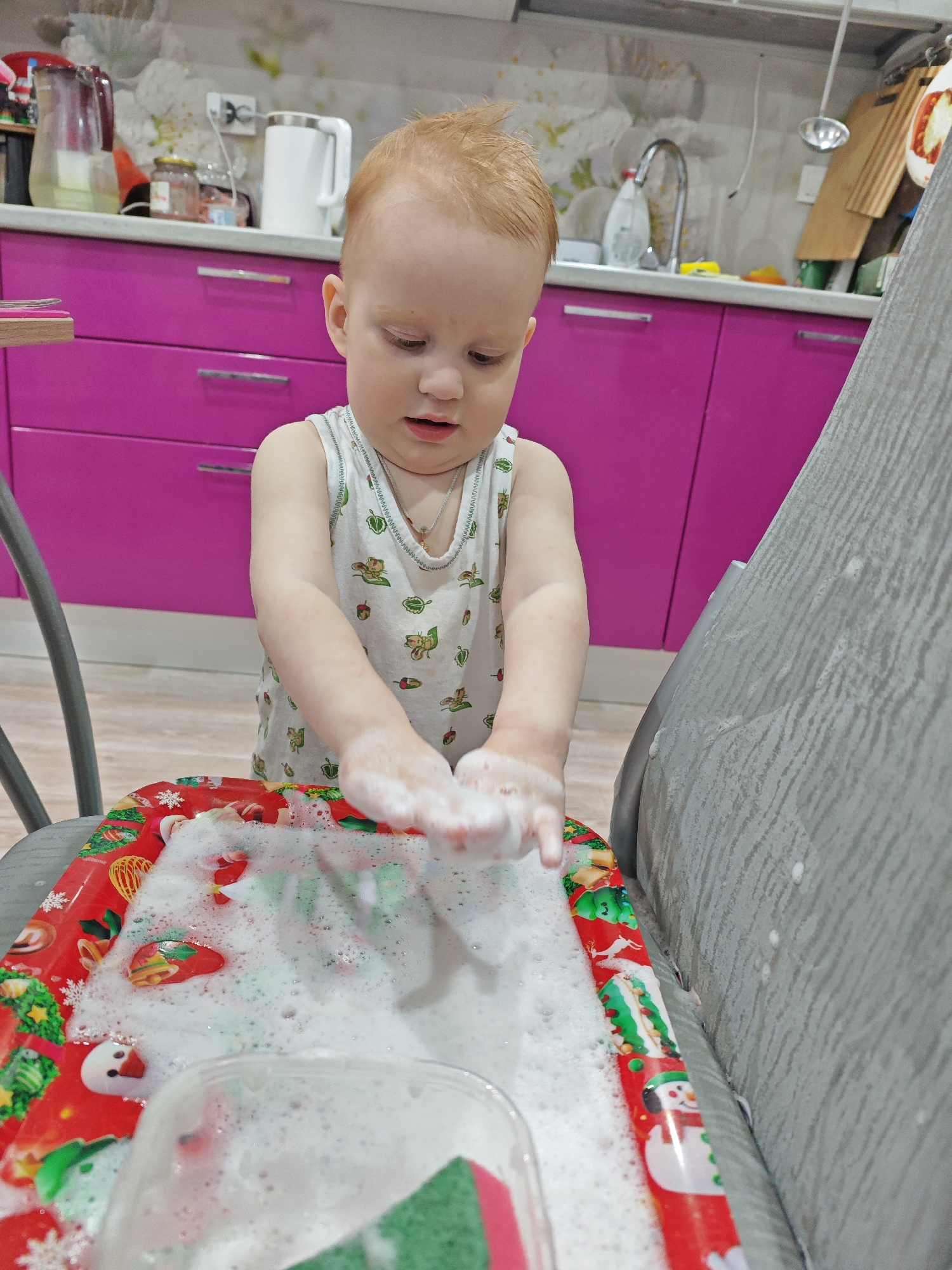 A young child with light red hair is depicted playing with foamy water in a kitchen setting in Markovskii, Russia. The child, appearing to be a toddler, wears a white sleeveless top patterned with small green designs, possibly leaves or acorns, and a thin silver chain necklace. Their hands are submerged in and covered with white suds, suggesting they are washing something or playing with bubbles. The child's gaze is directed downwards, focused intently on the activity in front of them.
The foamy water is contained within a red, rectangular tray adorned with festive Christmas decorations, including snowmen, snowflakes, Christmas trees, and bells. A green and red kitchen sponge is partially visible within a clear container inside the tray.
The background reveals a modern kitchen interior with eye-catching magenta lower cabinets featuring silver handles. A light-colored countertop holds various kitchen items such as a clear pitcher, a white electric kettle, and glass jars. Above the counter, a light-toned wall with a subtle, possibly floral or abstract, pattern is visible. A kitchen faucet and sink area can be seen in the distance. To the right, a textured grey piece of furniture, possibly a sofa or chair arm, shows splashes of water or foam. Another grey furniture piece is partially visible on the left. The scene conveys a calm, engaging, and playful activity taking place indoors during what appears to be daytime. No visible text is legible.
