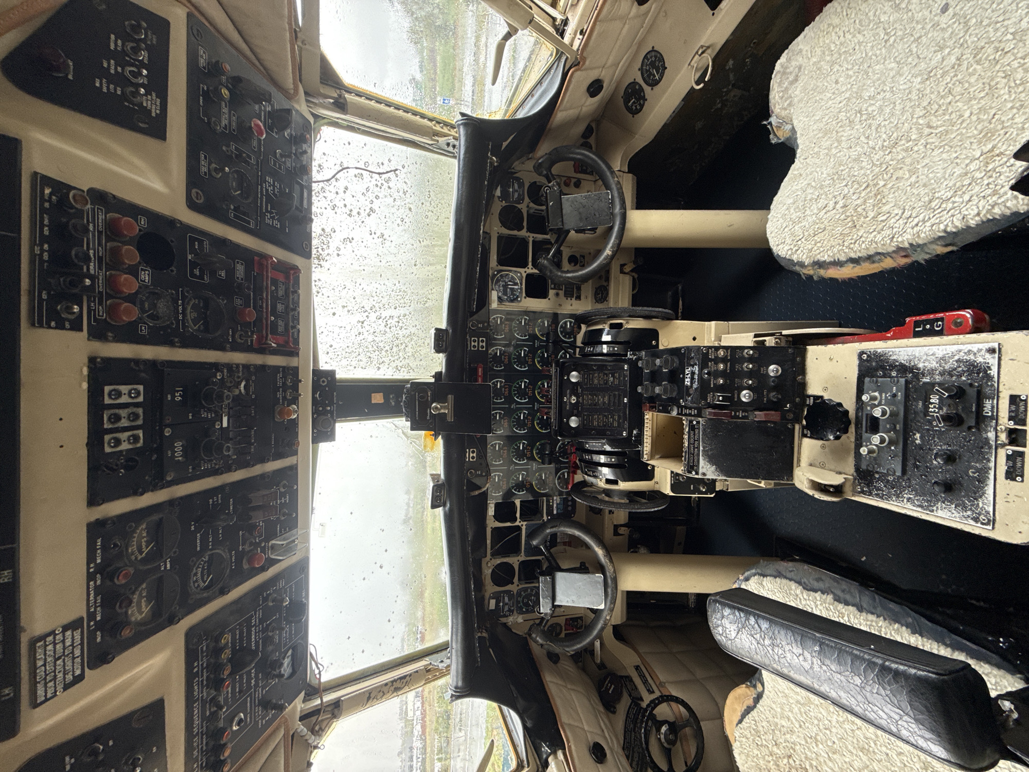 This is a photograph of the interior of an aircraft cockpit. The cockpit features a complex array of instruments, gauges, and control panels. Two pilot seats covered in a shaggy, light-colored material are visible. Through the front windshield, it is raining. The control panels display labels such as "LH ALTERNATOR," "RH MECH FAIL," "DE-ICE SEQUENCE LIGHTS RH," and "DME." The text "ZS-LYL" is also visible. There is no specific information available to pinpoint the location in Kraaifontein, South Africa. However, it can be implied to be at an aircraft museum or a similar facility.