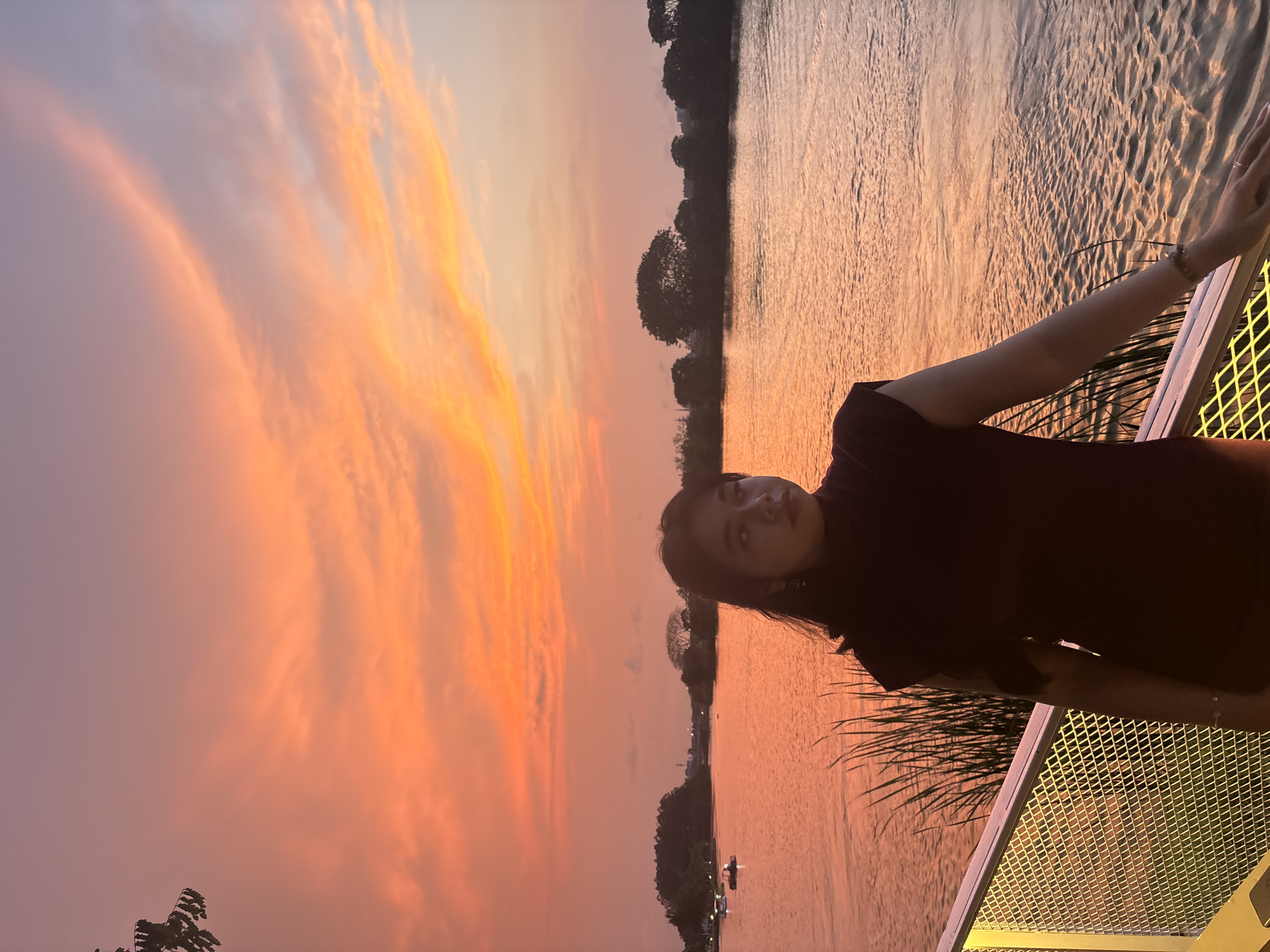 A woman is photographed at sunset on a riverfront in West Jakarta, Indonesia. She leans against a metal railing, gazing towards the camera. The sky is orange and pink. On the river, boats travel past trees.
