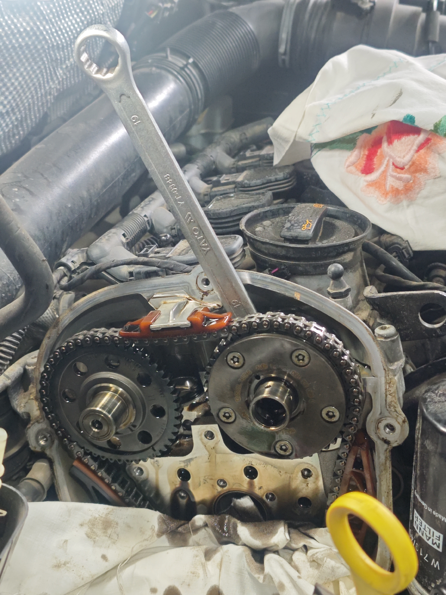 The image shows an open engine compartment of a vehicle under repair. A wrench labeled "19 YATO YT-0348" is resting on the engine's timing chain and gears. The timing chain and gears are exposed, with traces of oil visible. A white cloth with a red and orange floral pattern is draped nearby. A yellow dipstick handle is visible on the lower right and an oil filter is located next to it with the text "MANN FILTER Made in Germany."