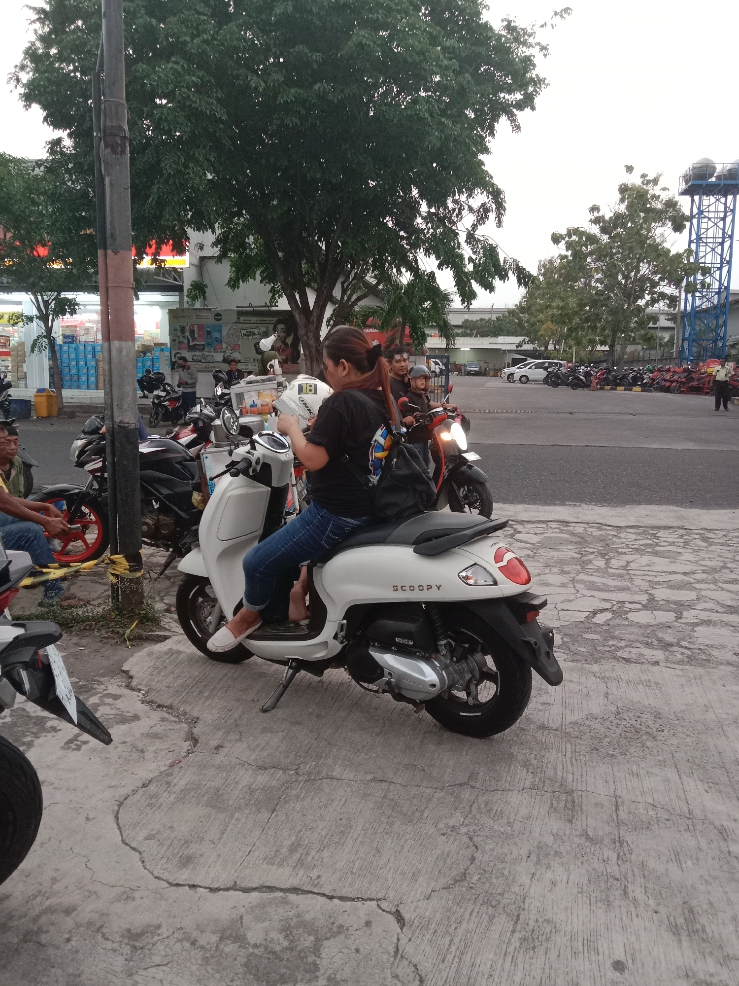 In Semarang, Indonesia, a woman sits on a white Honda Scoopy motorcycle, holding a white helmet with the number 18 on it. She is wearing blue jeans and white shoes. Behind her, another person is seated on a dark-colored motorcycle, also wearing a helmet. Several other motorcycles are parked in the background, along with a few cars and a tall blue water tower. The scene appears to be an outdoor parking area, possibly near a commercial establishment, with trees and a lamppost visible. The ground is paved with concrete.