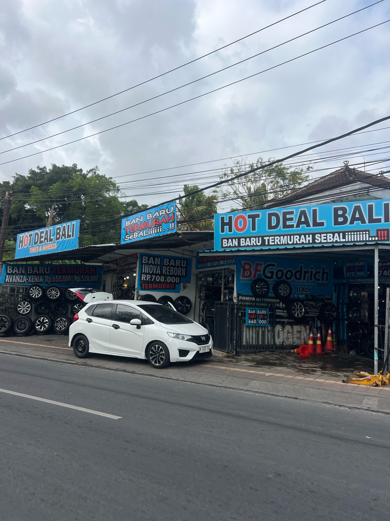 This is a street-level, eye-level, medium shot photograph taken on a cloudy day in Denpasar, Indonesia, showing a tire and wheel shop. The shop has several blue signs with white text. One prominent sign reads "HOT DEAL BALI TIRES & WHEELS". Another sign, partially obscured by a white car, states "BAN BARU TERMURAH SEBALiiiiii...!!". Below these signs, stacks of tires and wheels are displayed. A white hatchback car is parked in front of the shop, partially blocking the view of the tire displays. The car has a license plate that reads "N 3237 GV". On the right side of the shop, there is a sign with "BFGoodrich" in large letters. In front of the shop, there are several traffic cones and some yellow equipment, possibly for tire changing. The street in the foreground is asphalt with a white line marking. Trees are visible behind the shop, and telephone wires crisscross the sky above. The architecture of the buildings suggests a tropical urban environment.