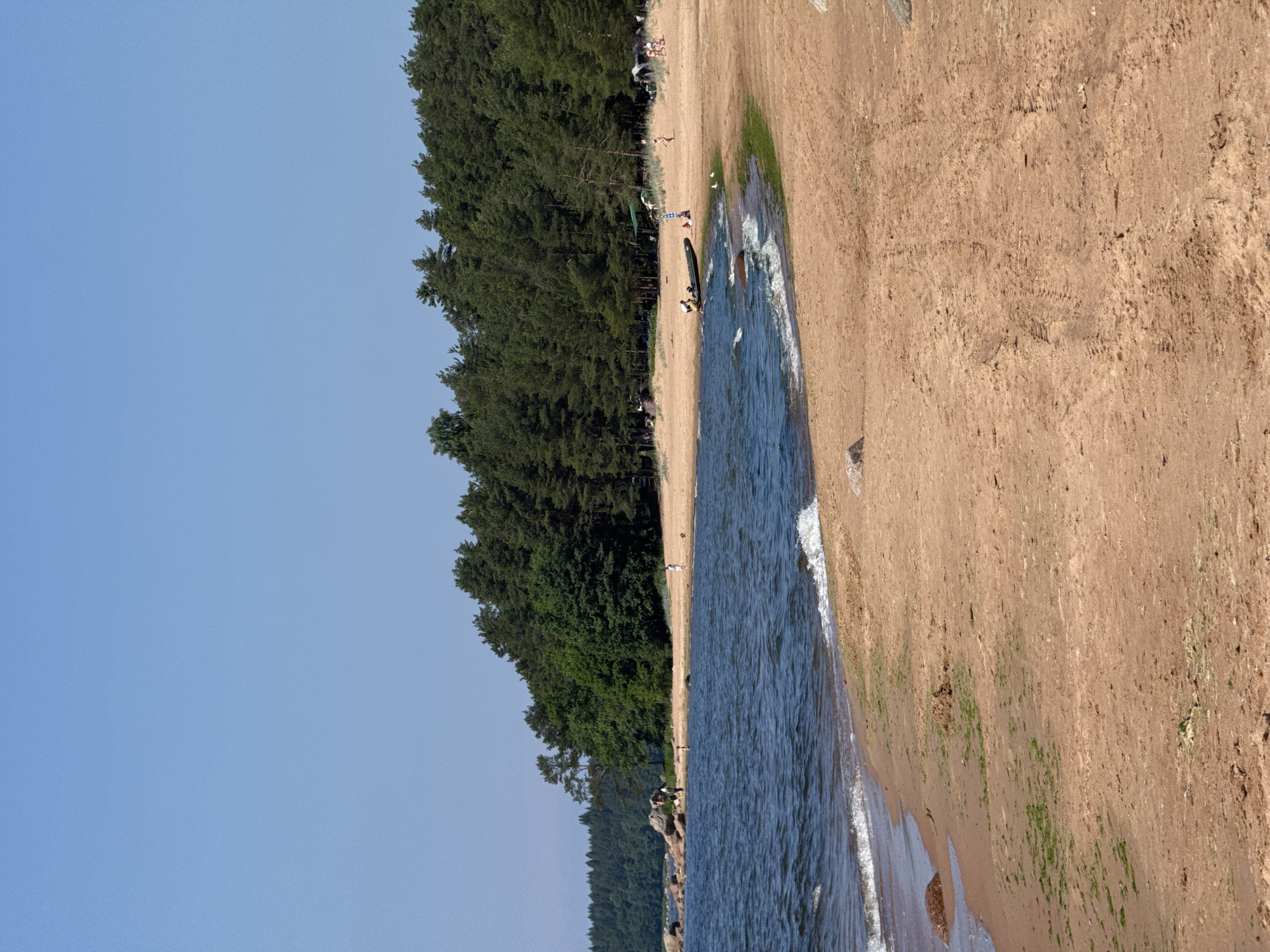 The image depicts a sandy beach with a body of dark blue water, likely a sea or large lake, in the foreground. Waves are gently lapping the shore, leaving a wet, darker sand line. The beach is dotted with some scattered green vegetation and rocks.
In the mid-ground, several people are present. On the left, near some large rocks, a few individuals are walking along the shore. Further to the right, a small inflatable boat is partially in the water, with a person near it. There are also other individuals scattered on the sand, some appearing to be walking or standing.
The background is dominated by a dense line of green pine trees, forming a forest that extends across the horizon. The sky above is a clear, bright blue with no clouds visible. The overall scene suggests a warm, sunny day at a natural beach location. The location is not specified to be in Zelenaya Roshcha, Russia.